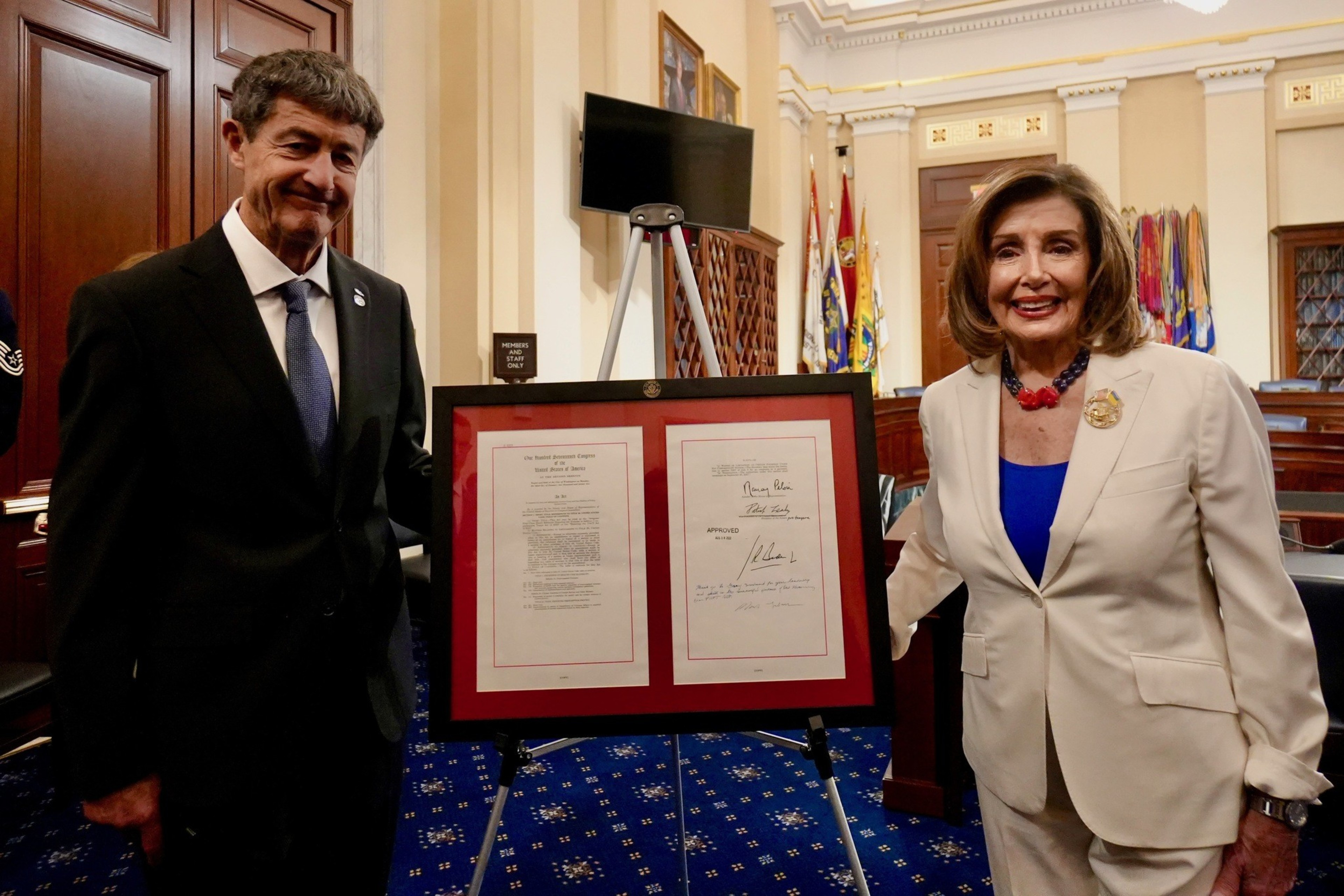 A man and a woman stand smiling beside two framed documents displayed on easels in a formal room with flags.