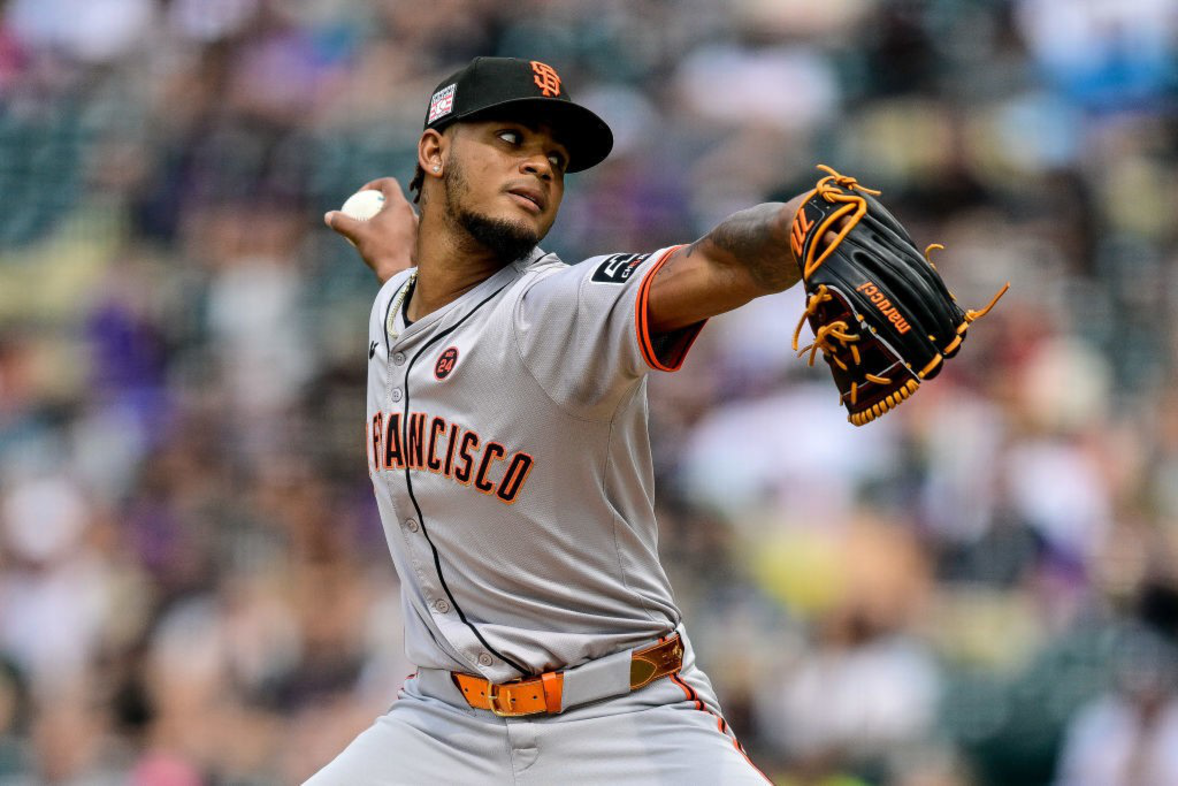 A baseball player in a gray uniform with "San Francisco" across the chest is pitching, wearing a black cap and glove, with a blurred stadium background.