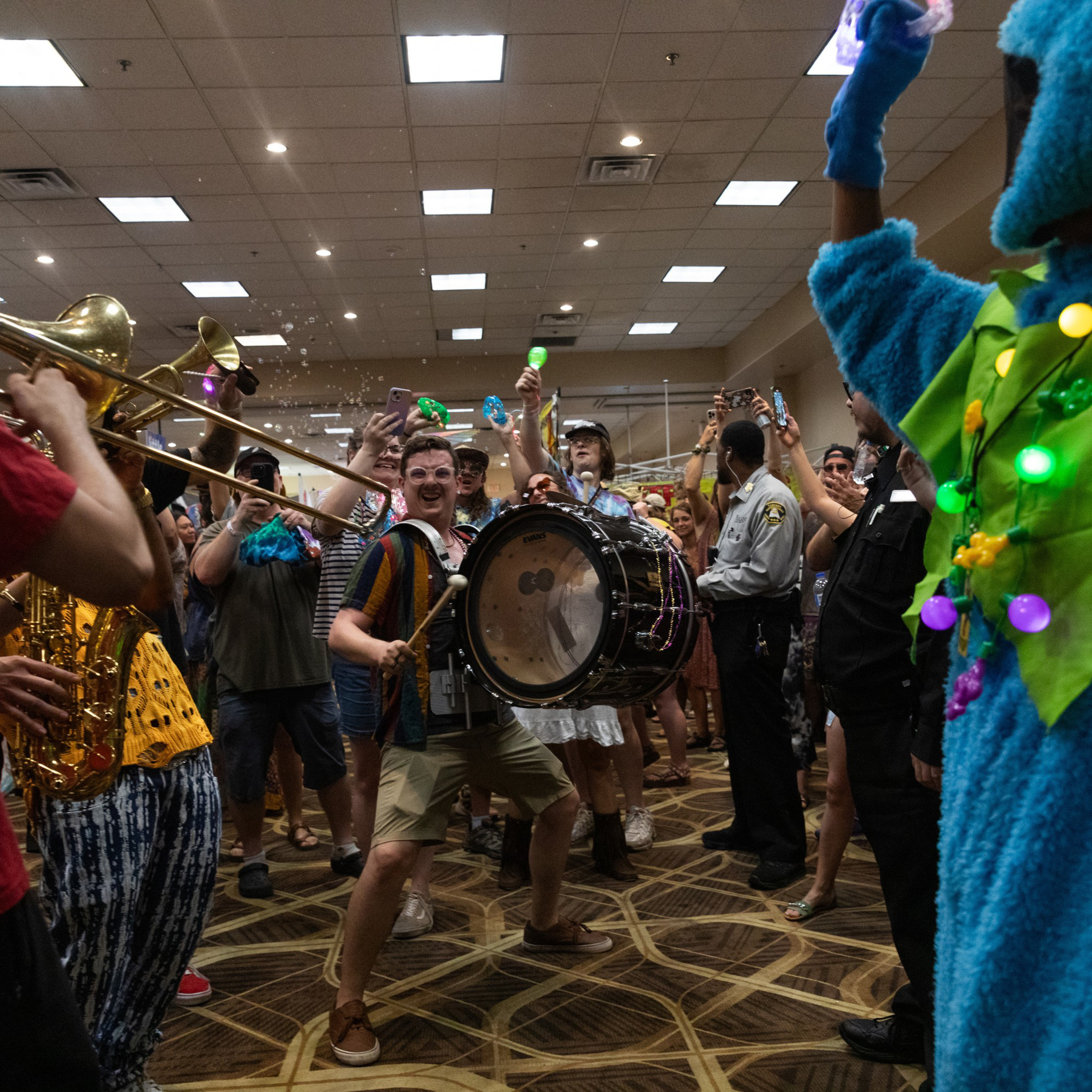 A lively crowd at an indoor event features musicians playing drums and brass instruments. Attendees, some in costumes, cheer, take photos, and blow bubbles.
