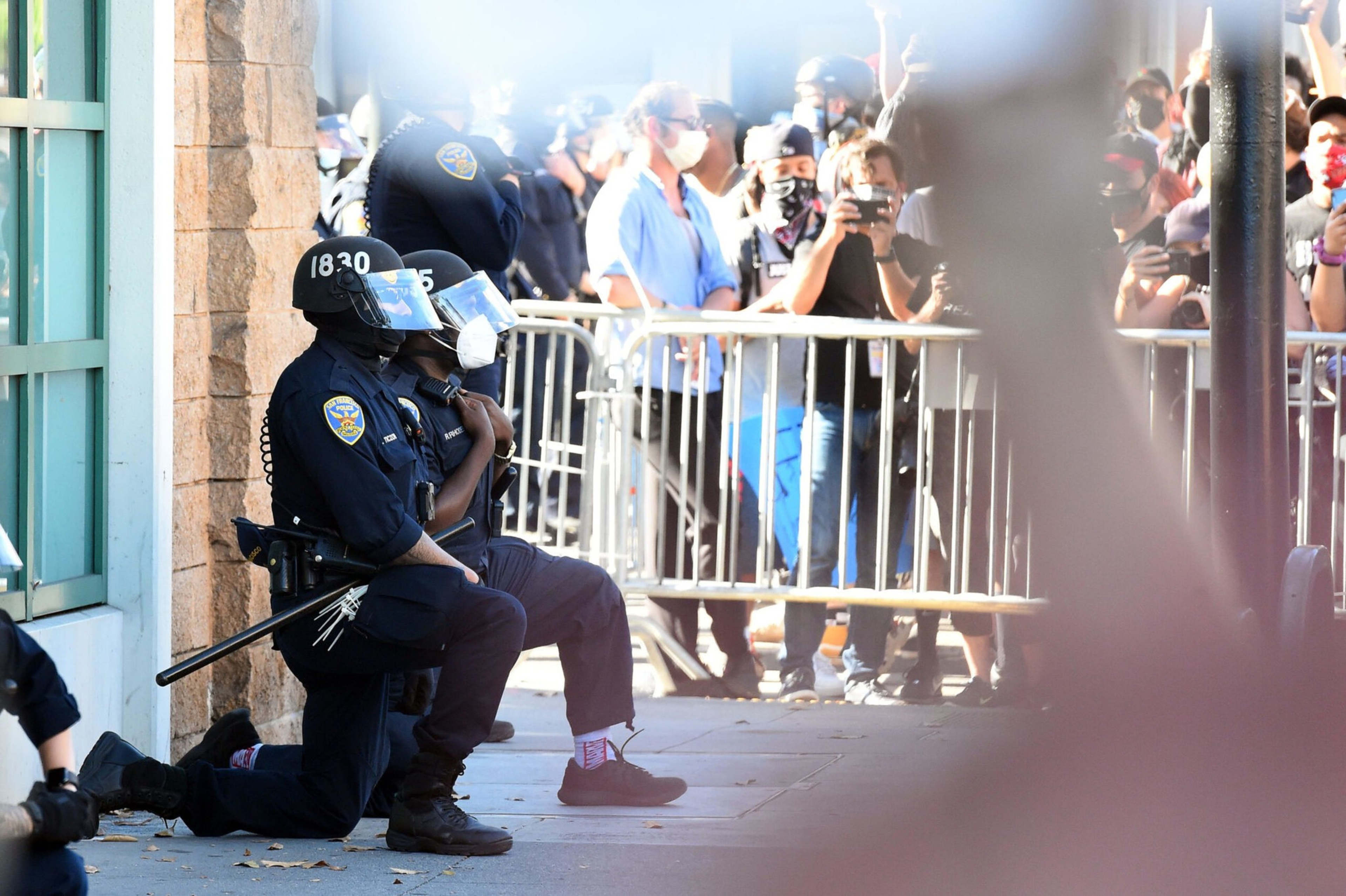 Two police officers wearing helmets and face shields kneel on the sidewalk, facing a crowd of masked protesters behind metal barricades.