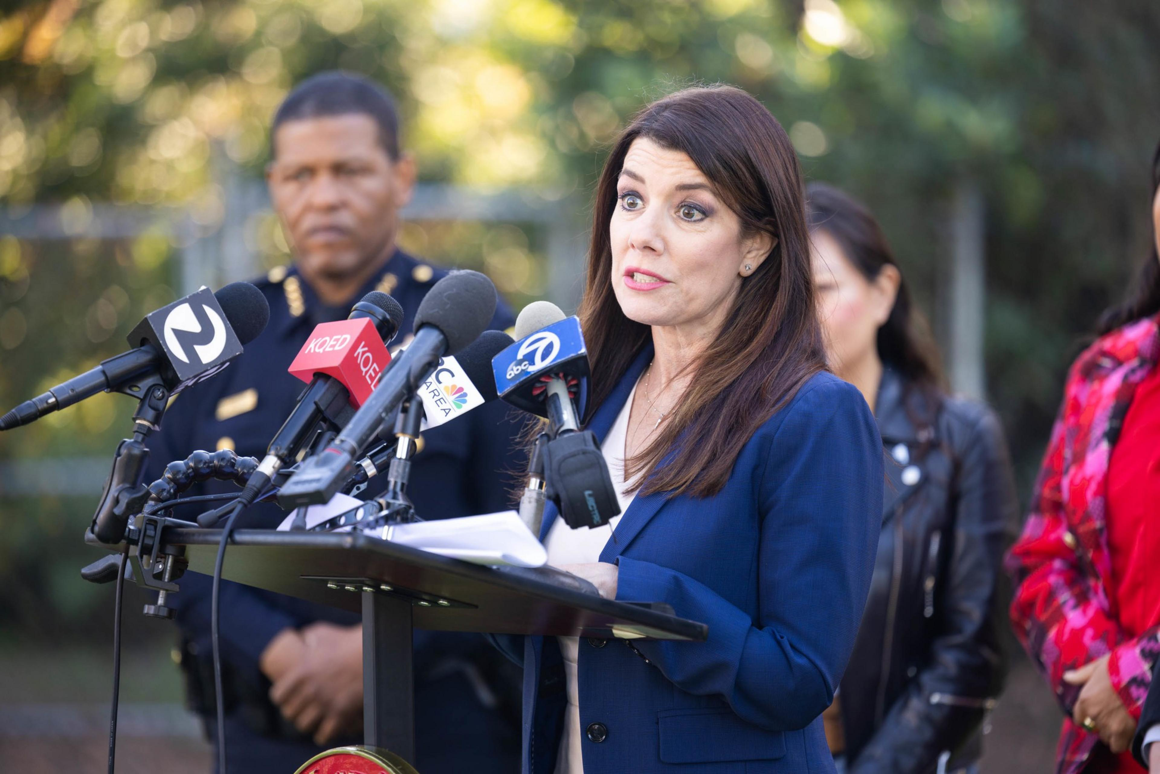 A woman in a blue blazer speaks at a podium surrounded by microphones from news outlets, with a man in uniform blurred in the background.
