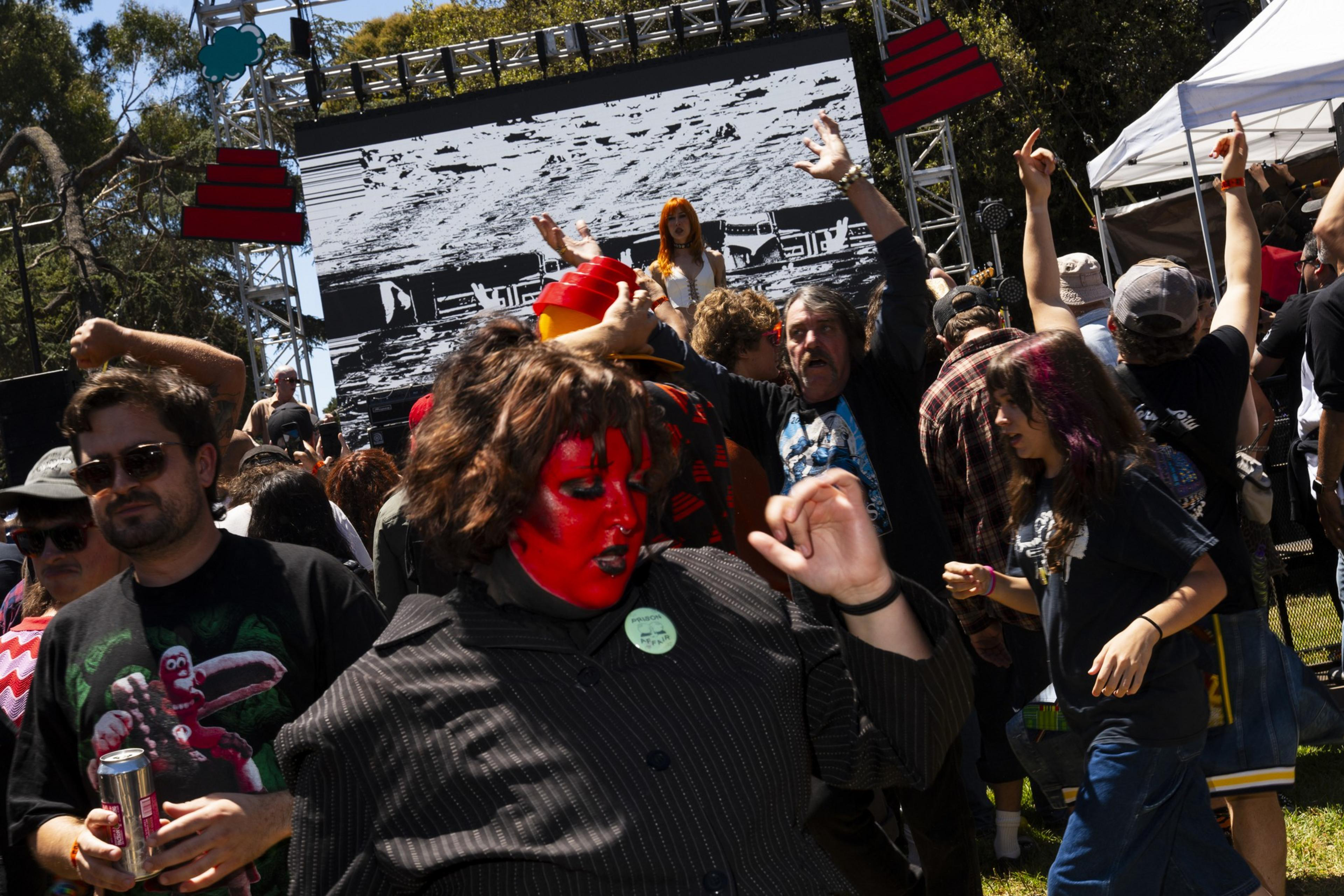 A lively outdoor scene with people dancing and celebrating. A person with a red painted face stands out in front of a stage with a large screen, under daylight.