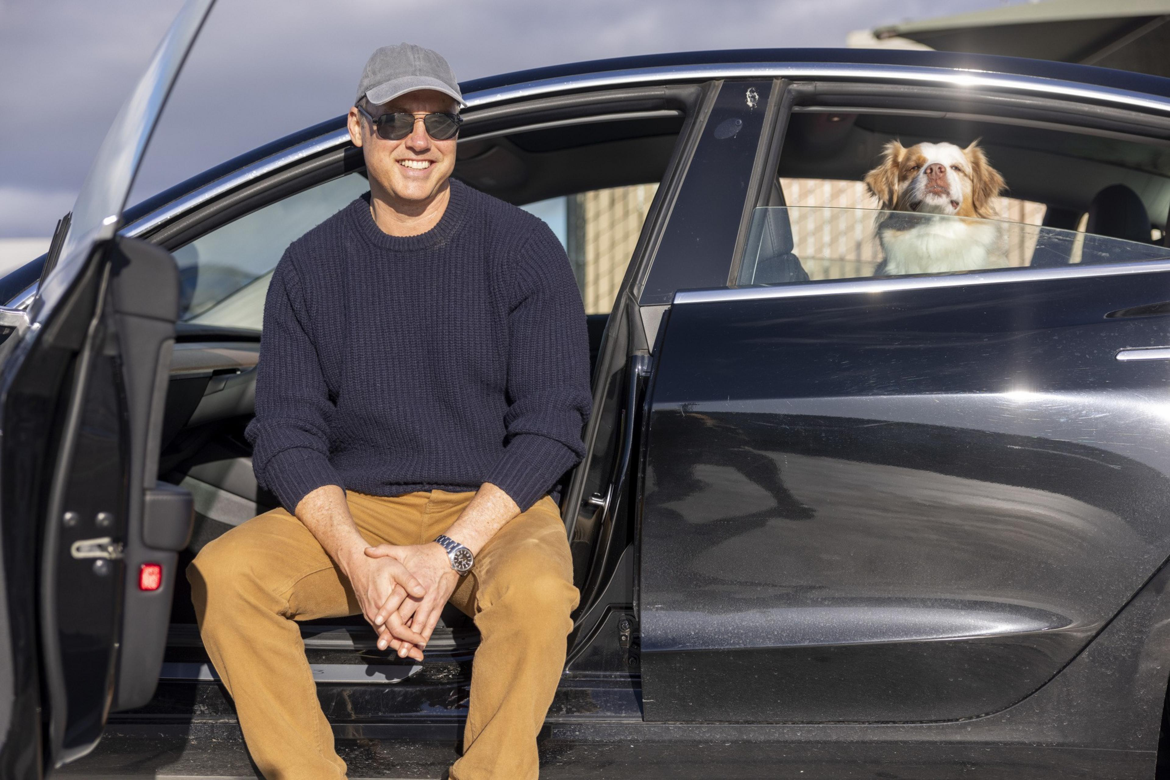 A man in a cap and sunglasses sits on a black car's open door ledge, smiling. A dog is sitting inside the car, looking out the window.