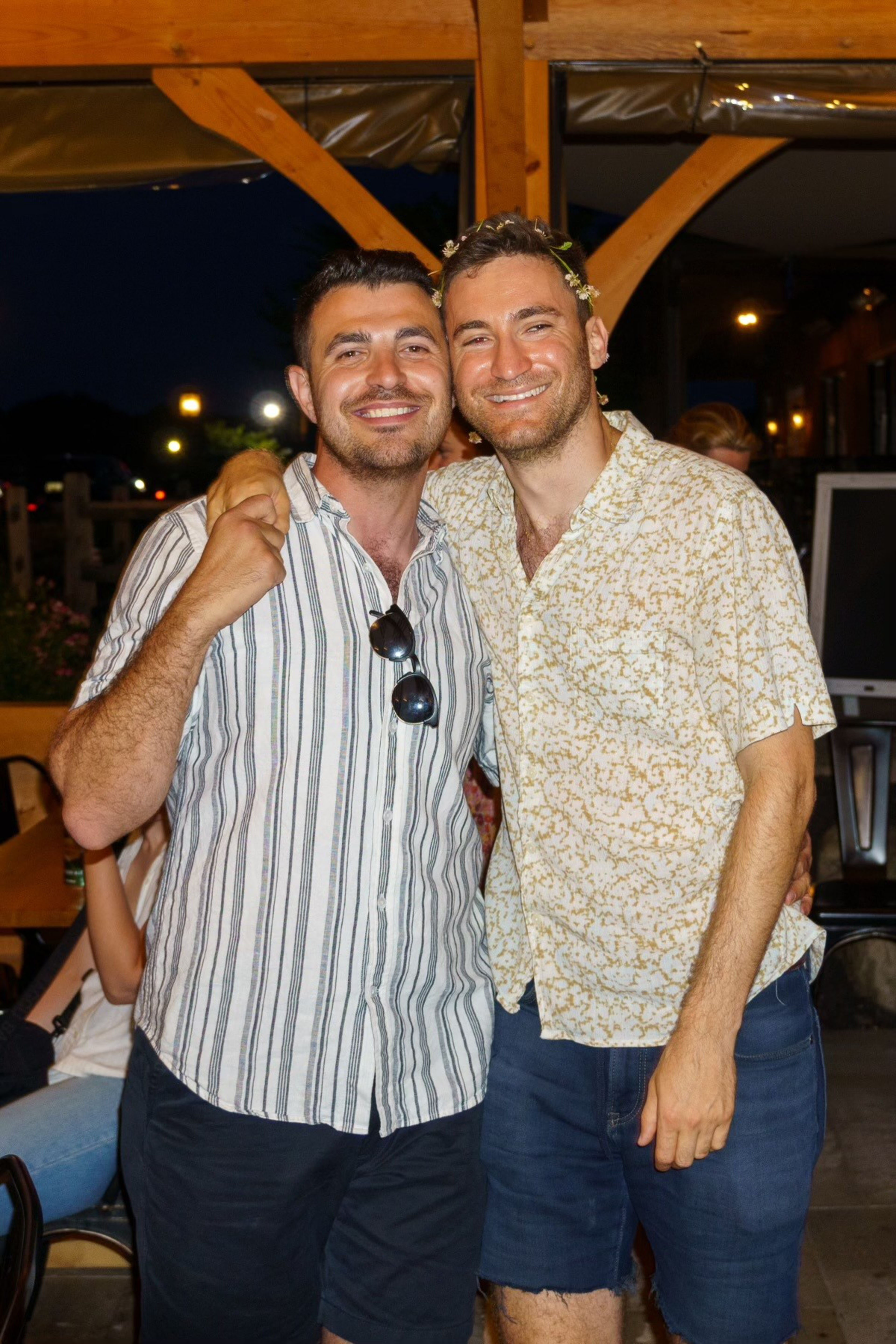 Two men smiling under a wooden pavilion at night, each wearing a patterned shirt and shorts. One has a floral crown, and they are standing close together.