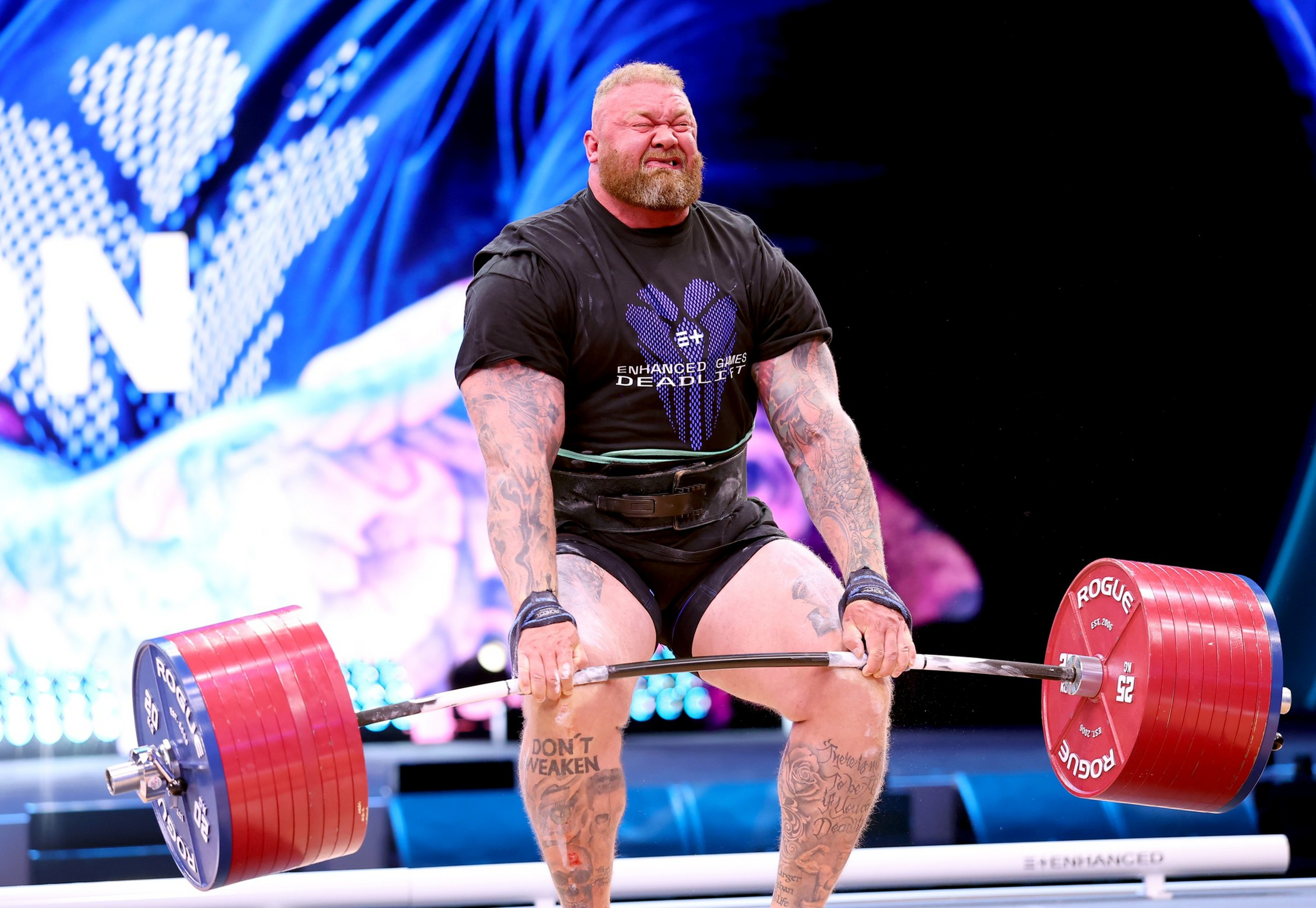 A muscular man with tattoos strains while deadlifting a heavily weighted barbell during a competition.