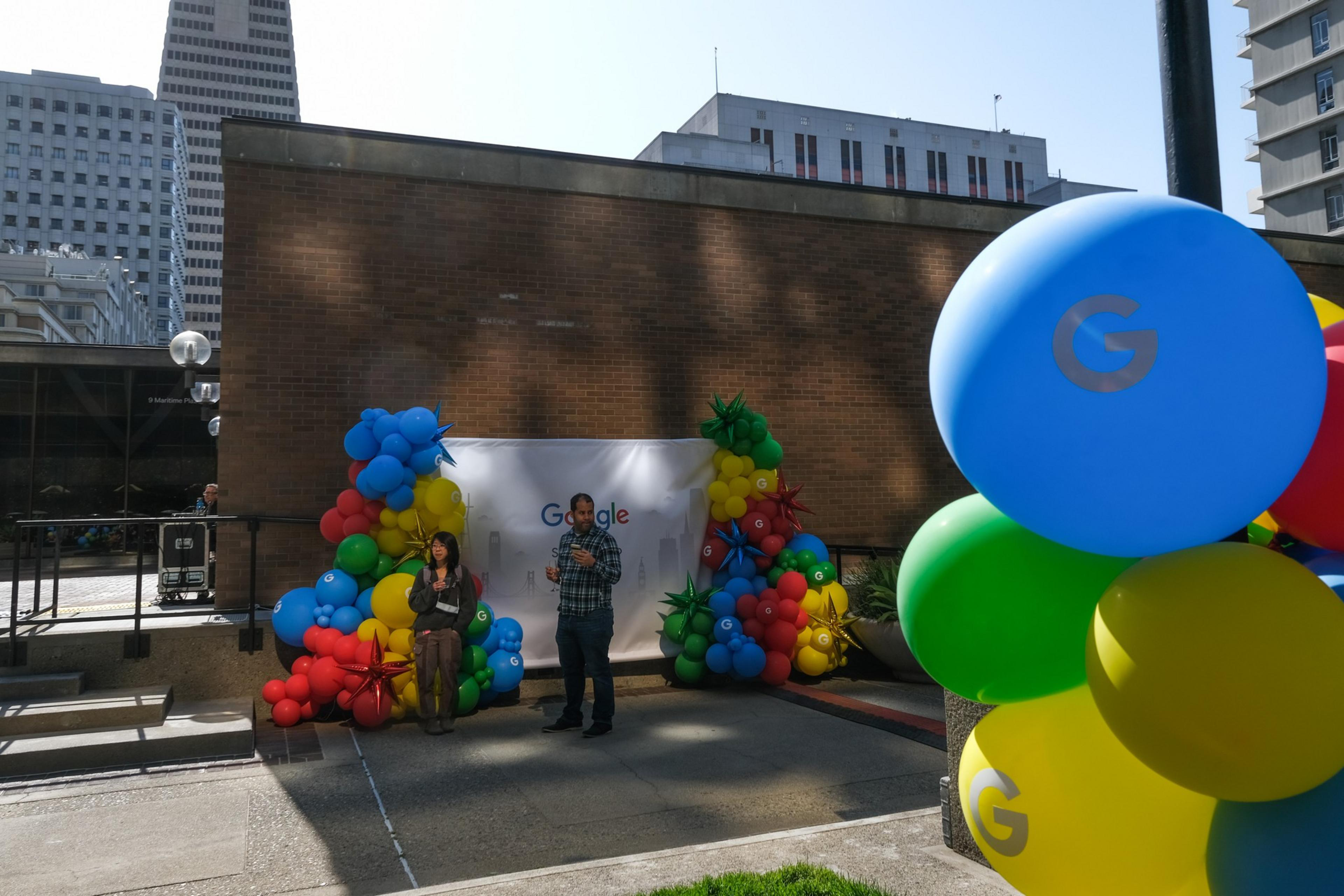 Two people stand near a colorful balloon arch with Google branding, set against an urban backdrop with tall buildings and a brick wall.