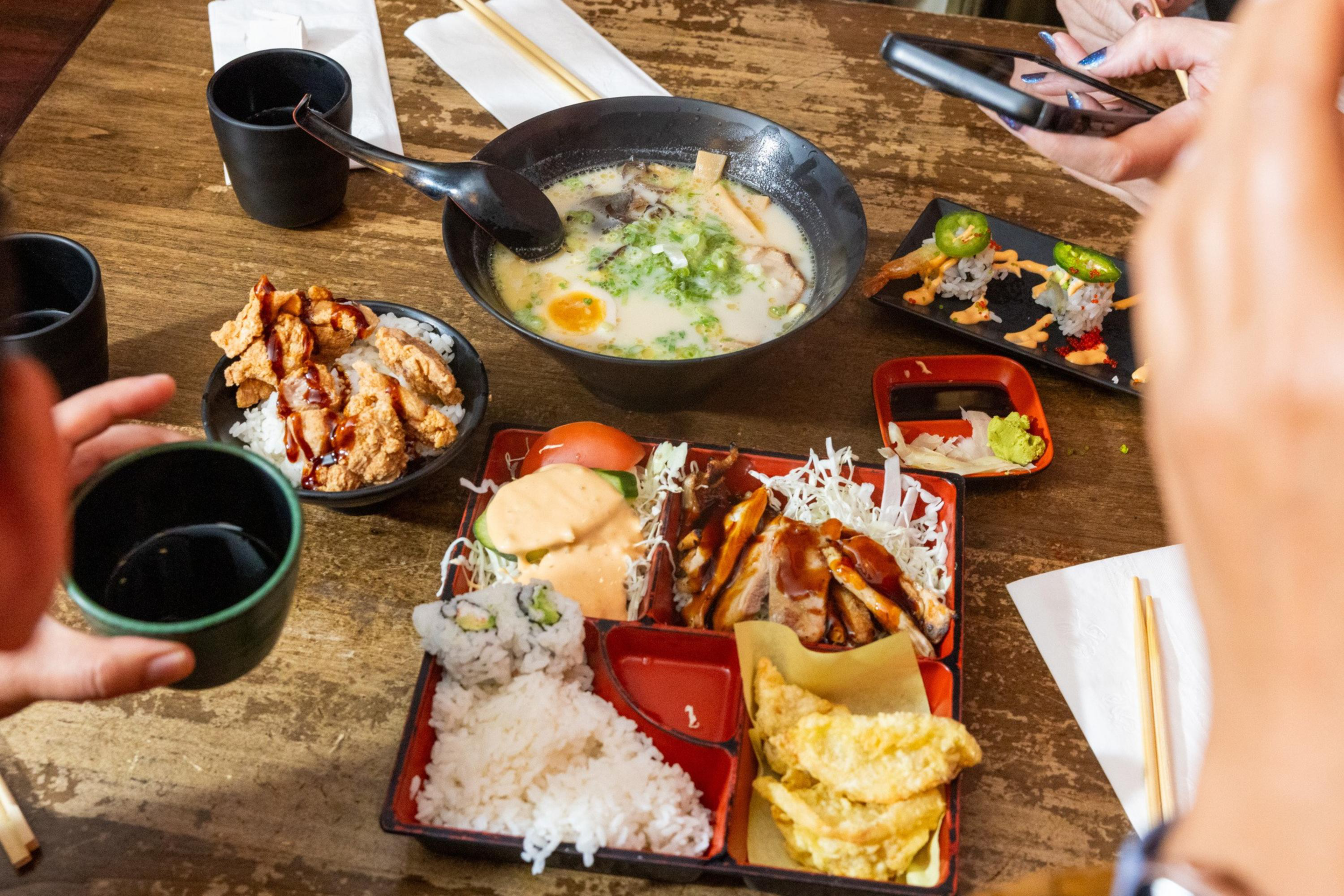A table with bowls of ramen, fried chicken, sushi rolls, a bento box with rice, grilled meat, tempura, and two people holding cups and a phone.