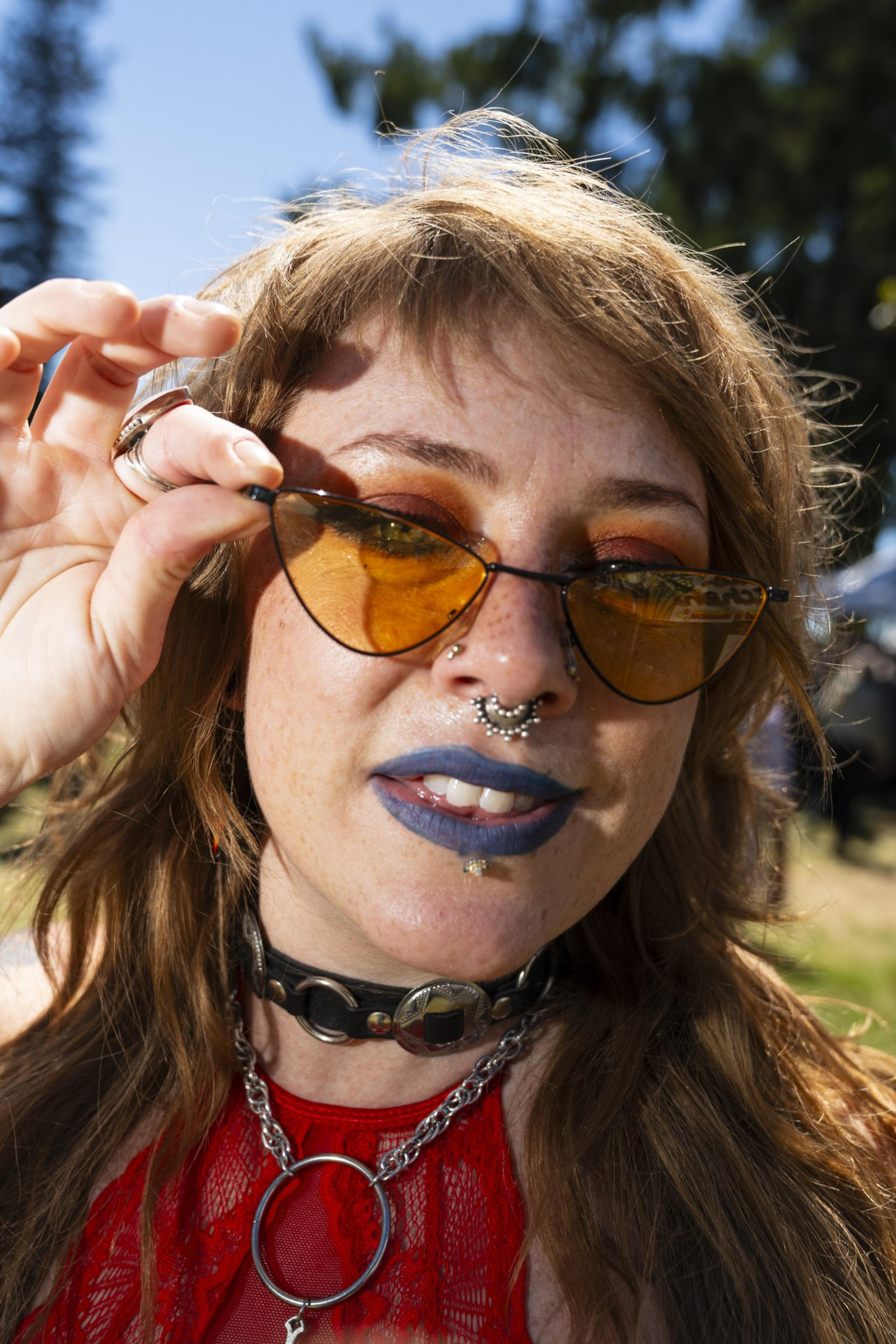 A person with blue lipstick and septum piercing touches orange cat-eye sunglasses, wearing a black choker and red lace top, outdoors.