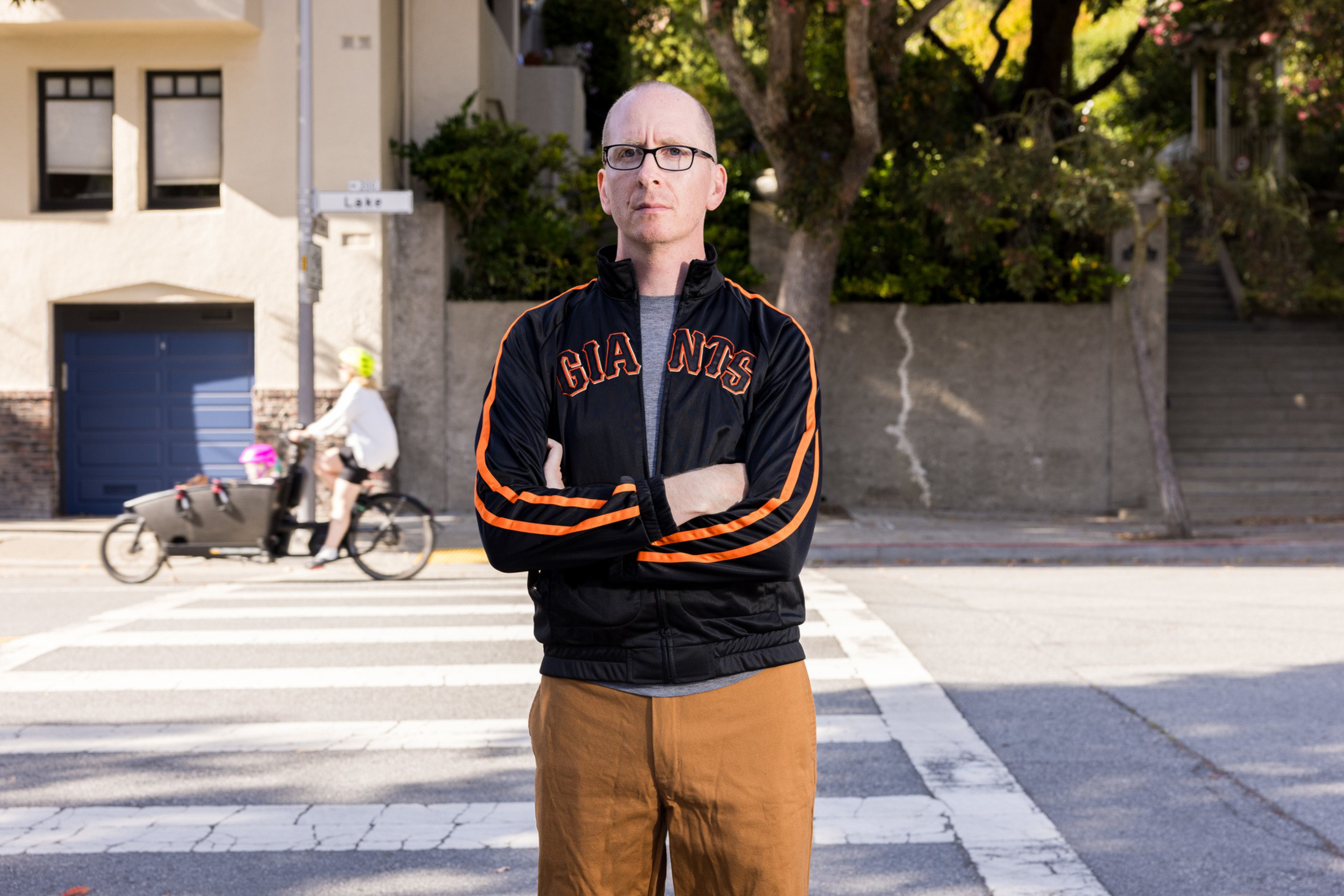 A man with glasses wearing a black Giants jacket and brown pants stands with arms crossed on a city crosswalk.