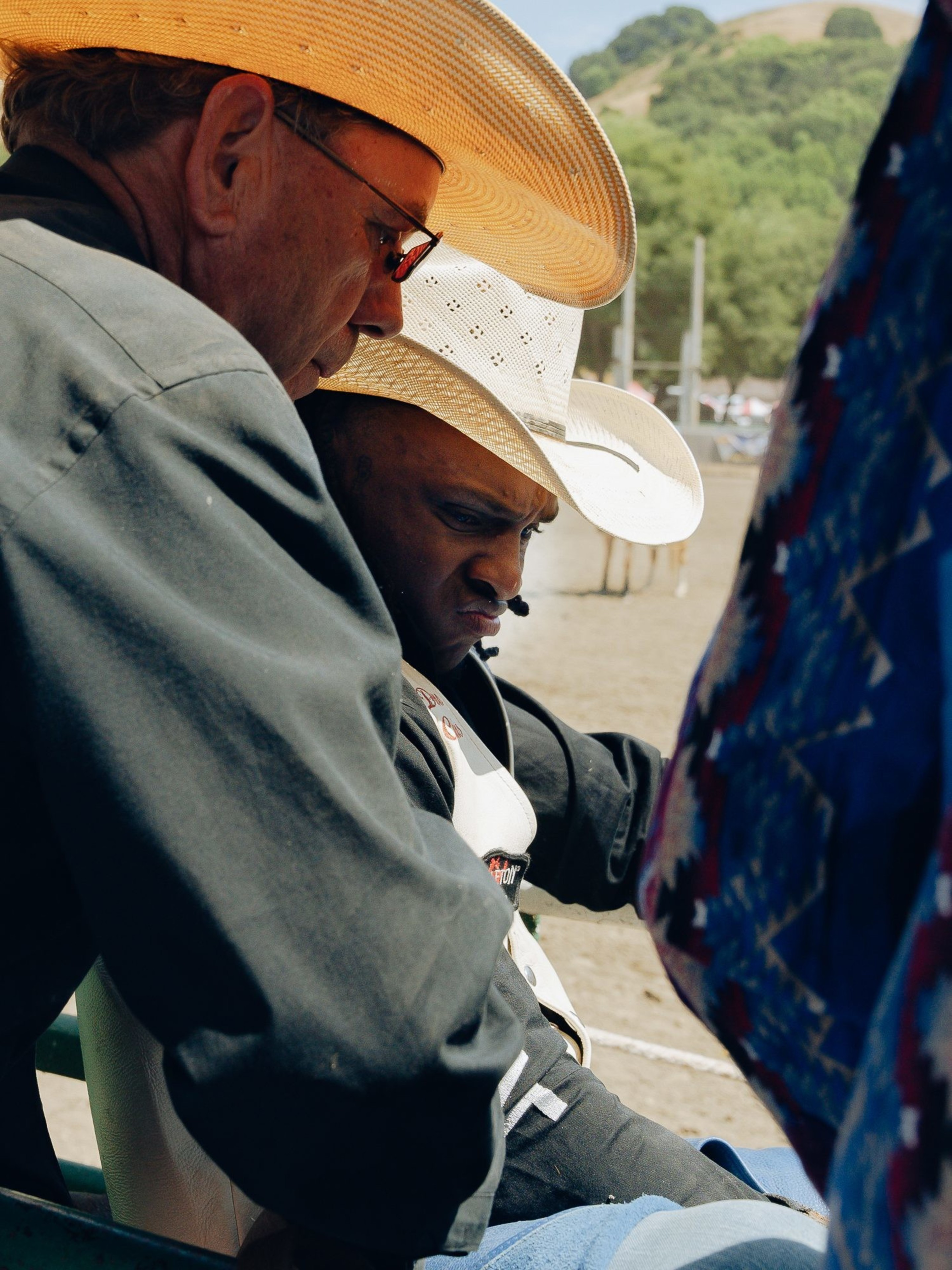 Two men, both wearing large cowboy hats, are engaged in conversation outdoors. One man is sitting and appears focused, while the other leans in closely. Trees and a hill are in the background.
