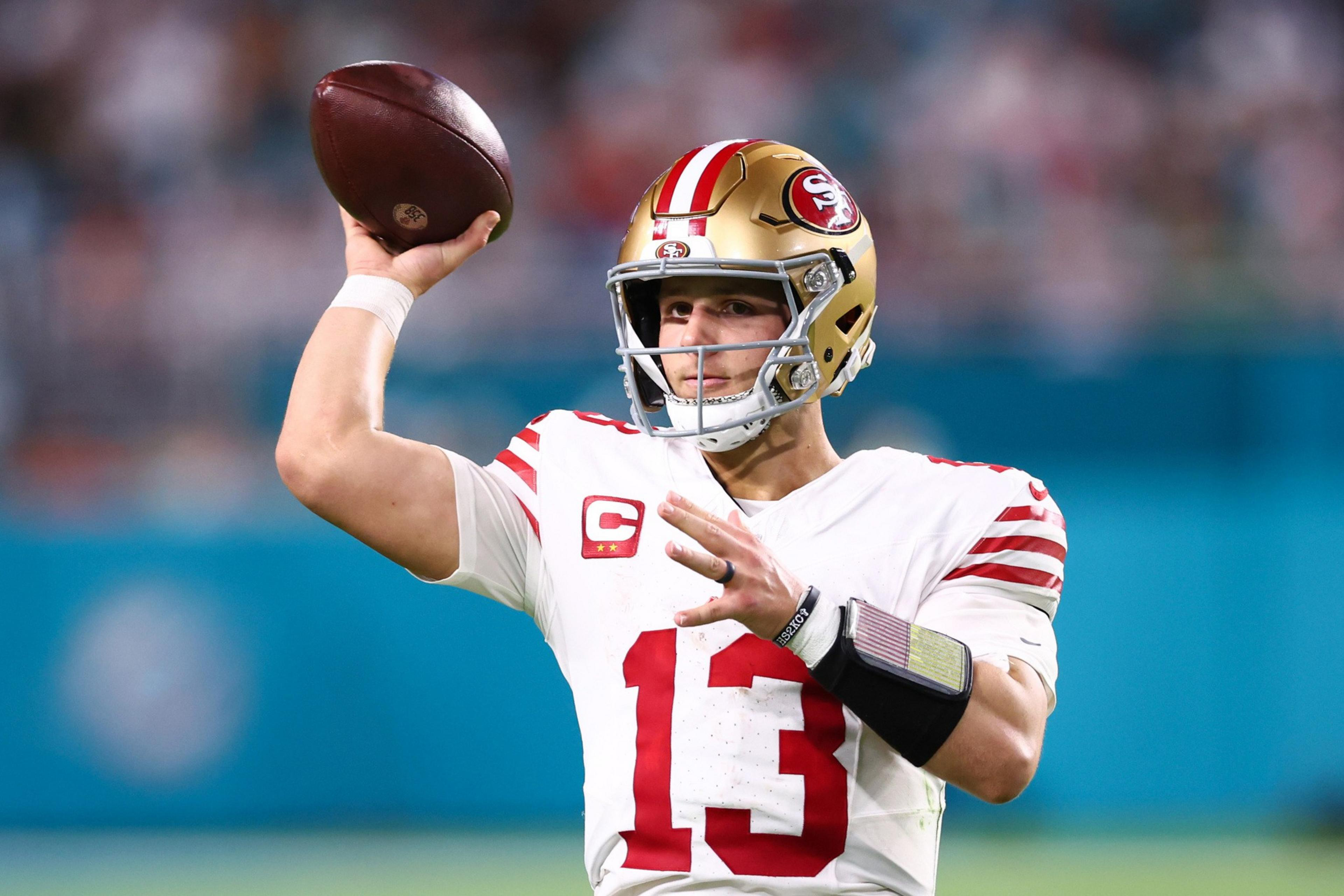 A football player in a white and red uniform prepares to throw a pass. He wears a golden helmet and holds a football in his right hand, aiming downfield.