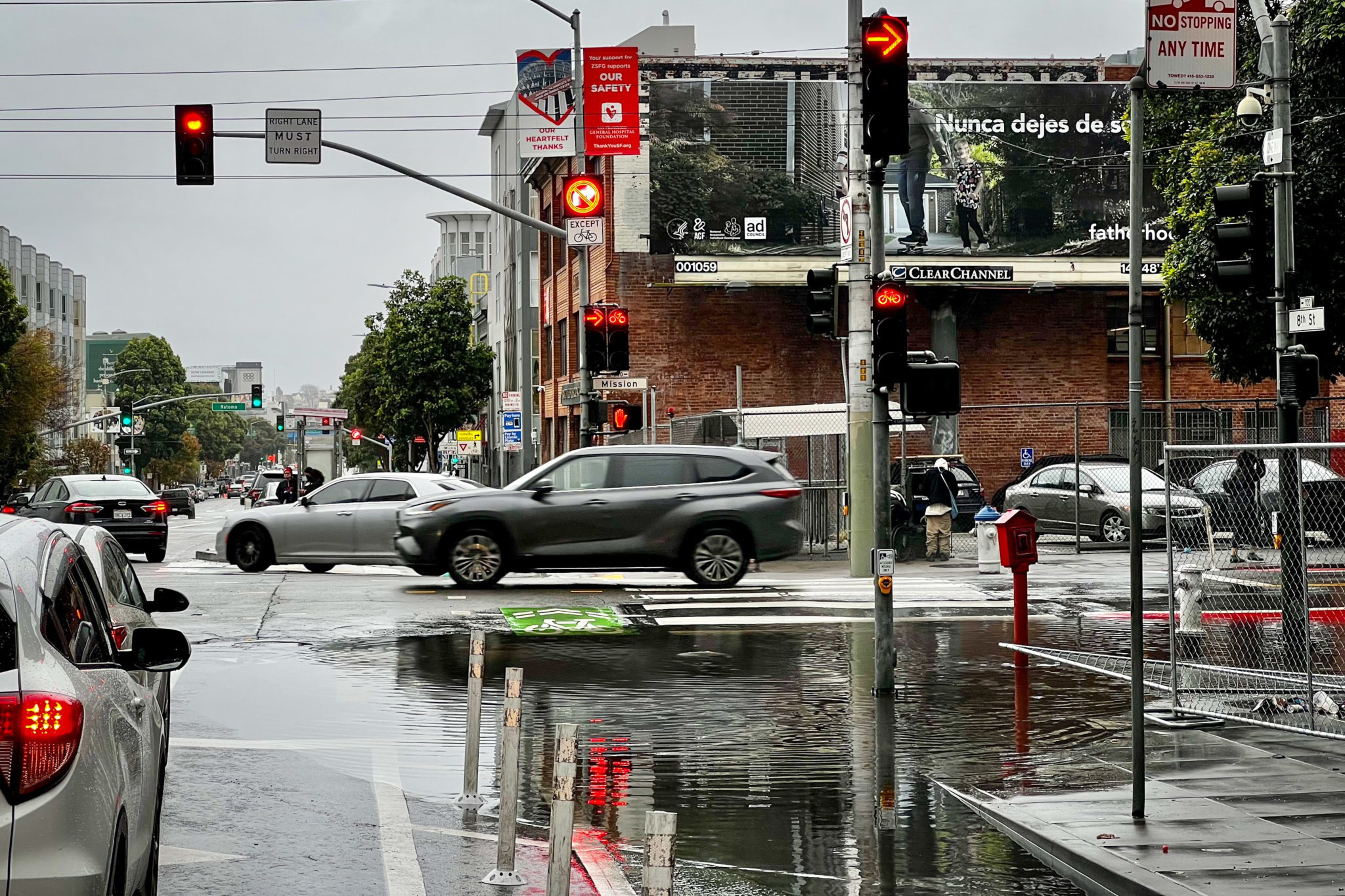 A wet city intersection with cars stopped at red lights, reflections on the wet pavement, and a large billboard in the background.