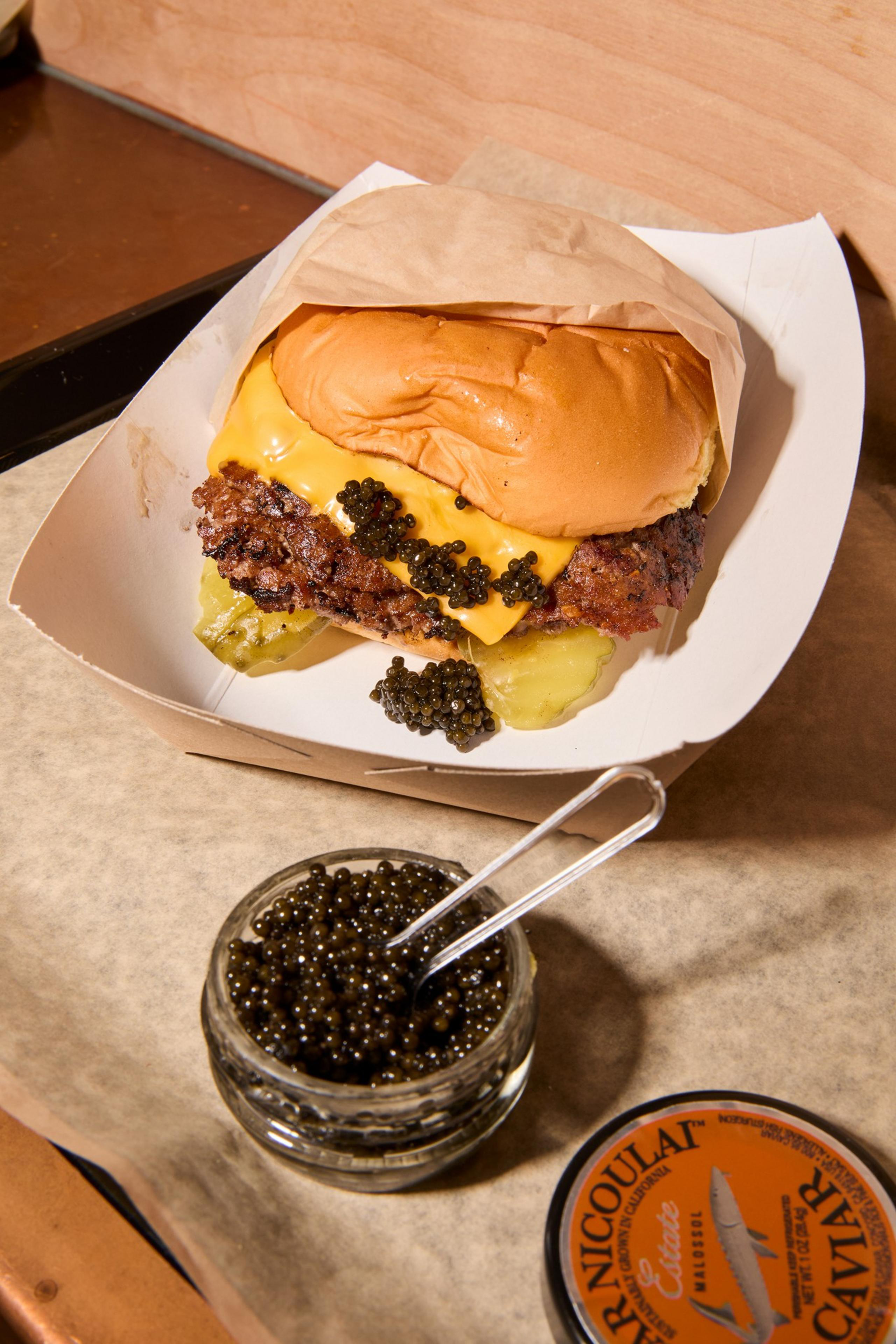 A cheeseburger with pickles and caviar on top sits in a paper tray. Beside it, there's an open jar of caviar with a small spoon.