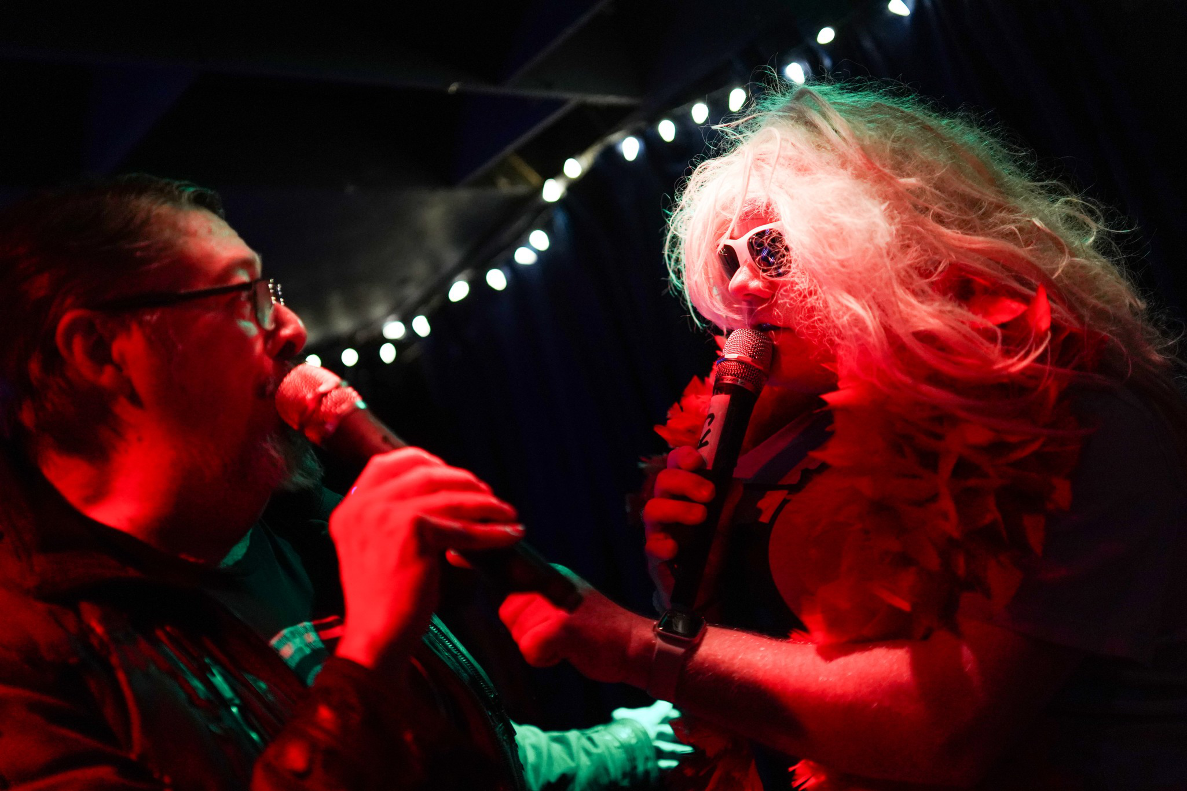 Two people sing into microphones on stage, one wearing glasses and a leather jacket, the other in a blonde wig, sunglasses, and a red feather boa.