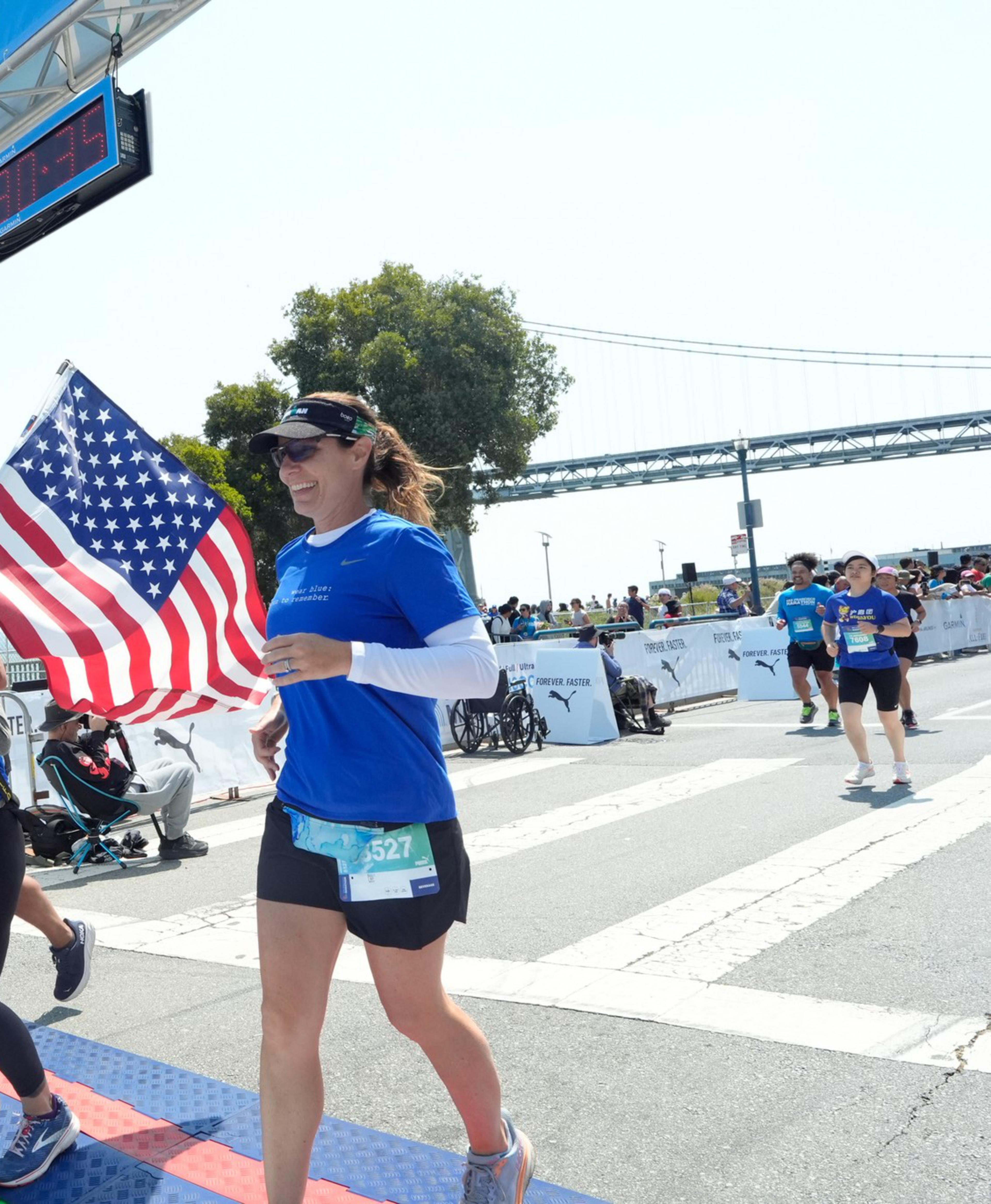 Two women in blue shirts, one holding an American flag, are crossing the finish line of a marathon, with a bridge and spectators in the background.