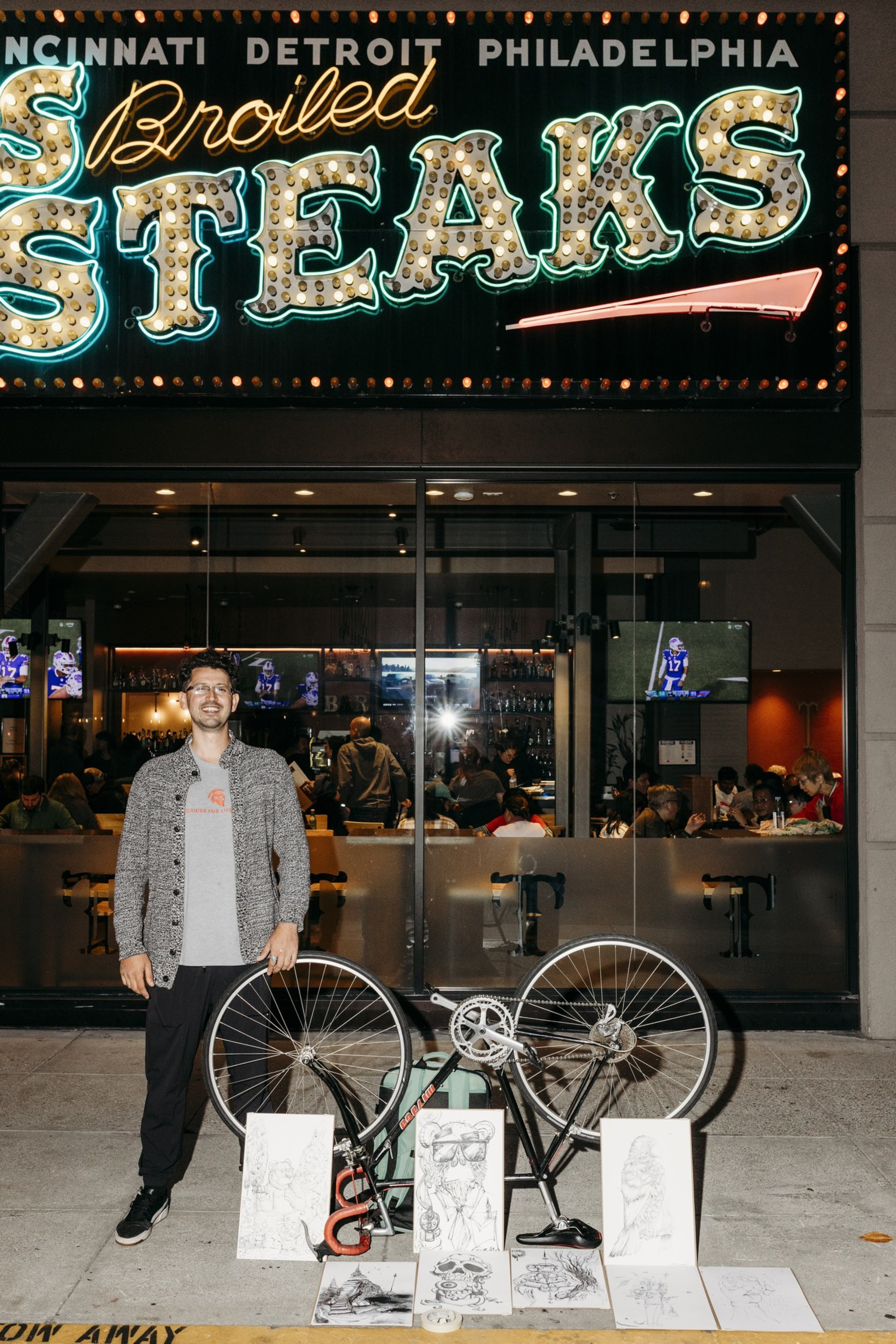 A man stands outside a steakhouse with a bicycle and several black-and-white sketches displayed on the ground in front of him.