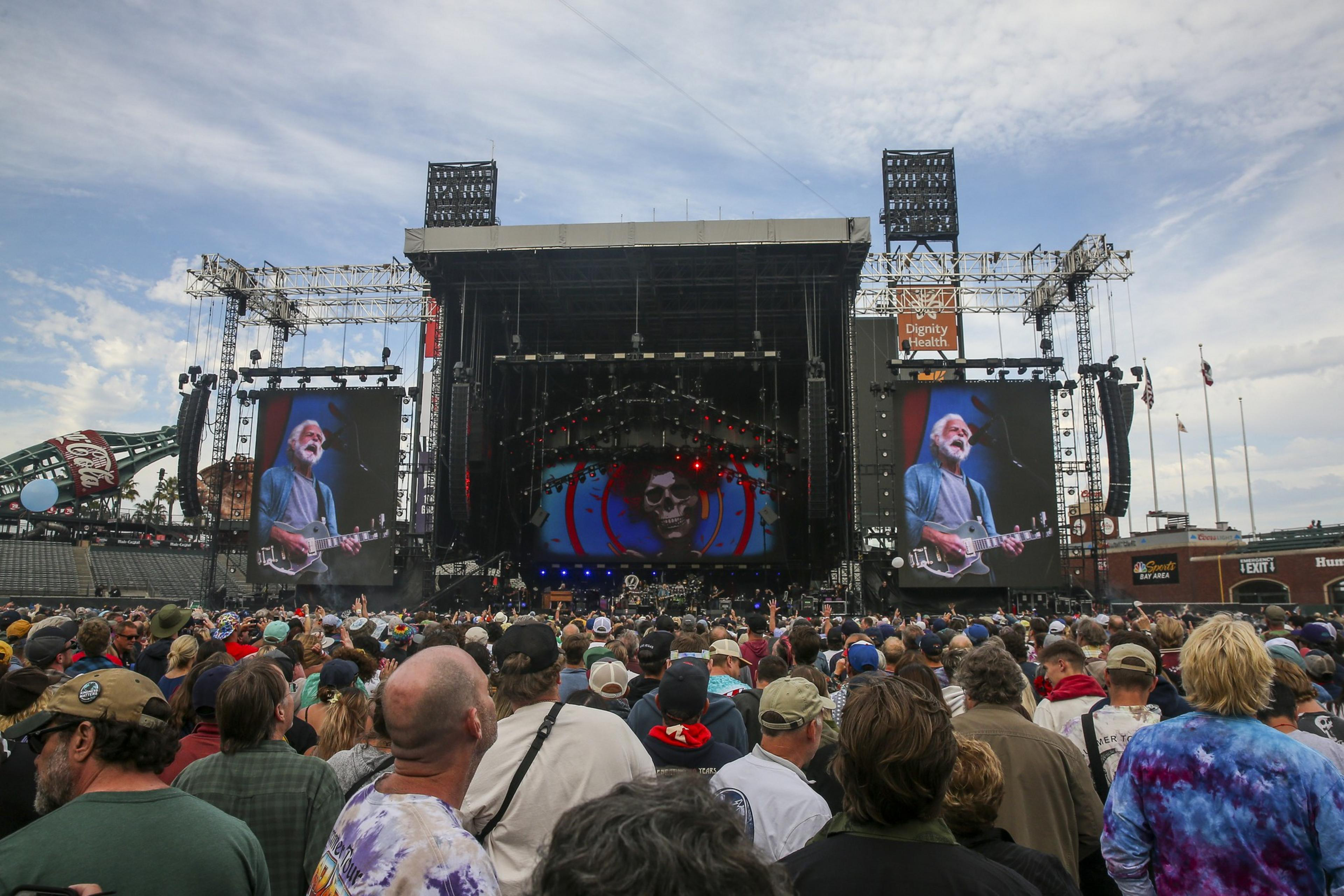 A large crowd faces a stage at a concert. Screens display a musician playing guitar. The background features colorful lights and a skull image.