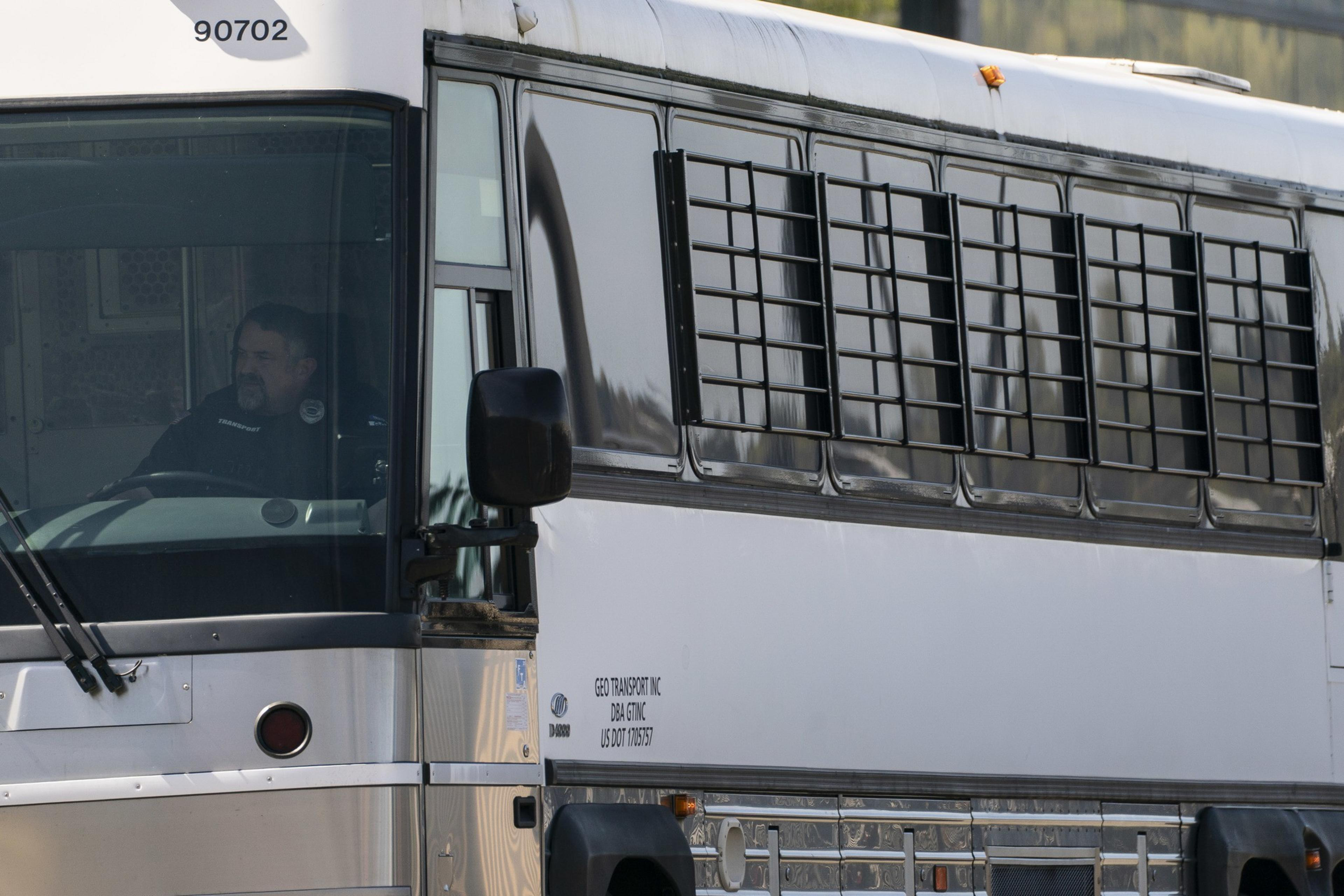 A bus with barred windows is driven by a person in a uniform. The bus is labeled &quot;GEO Transport Inc.&quot; and has vehicle numbers visible.