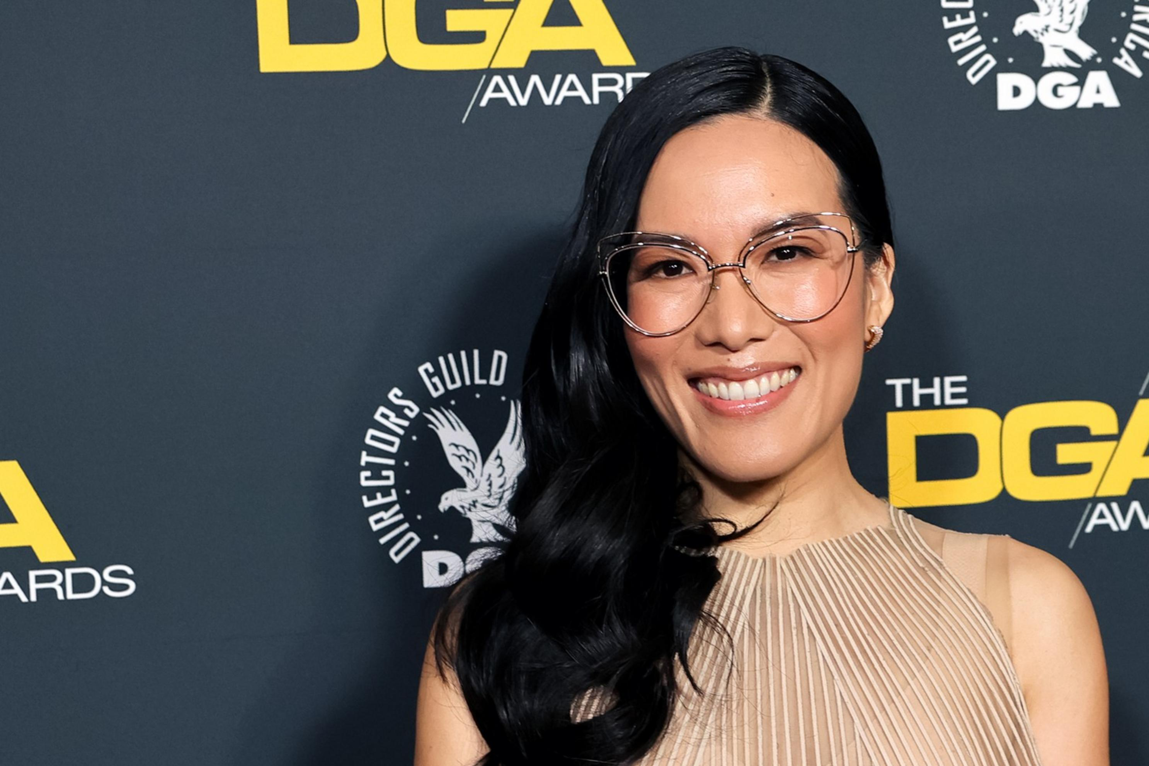 A person with long dark hair, wearing glasses and a beige dress, smiles against a backdrop featuring yellow "DGA Awards" text and logos.