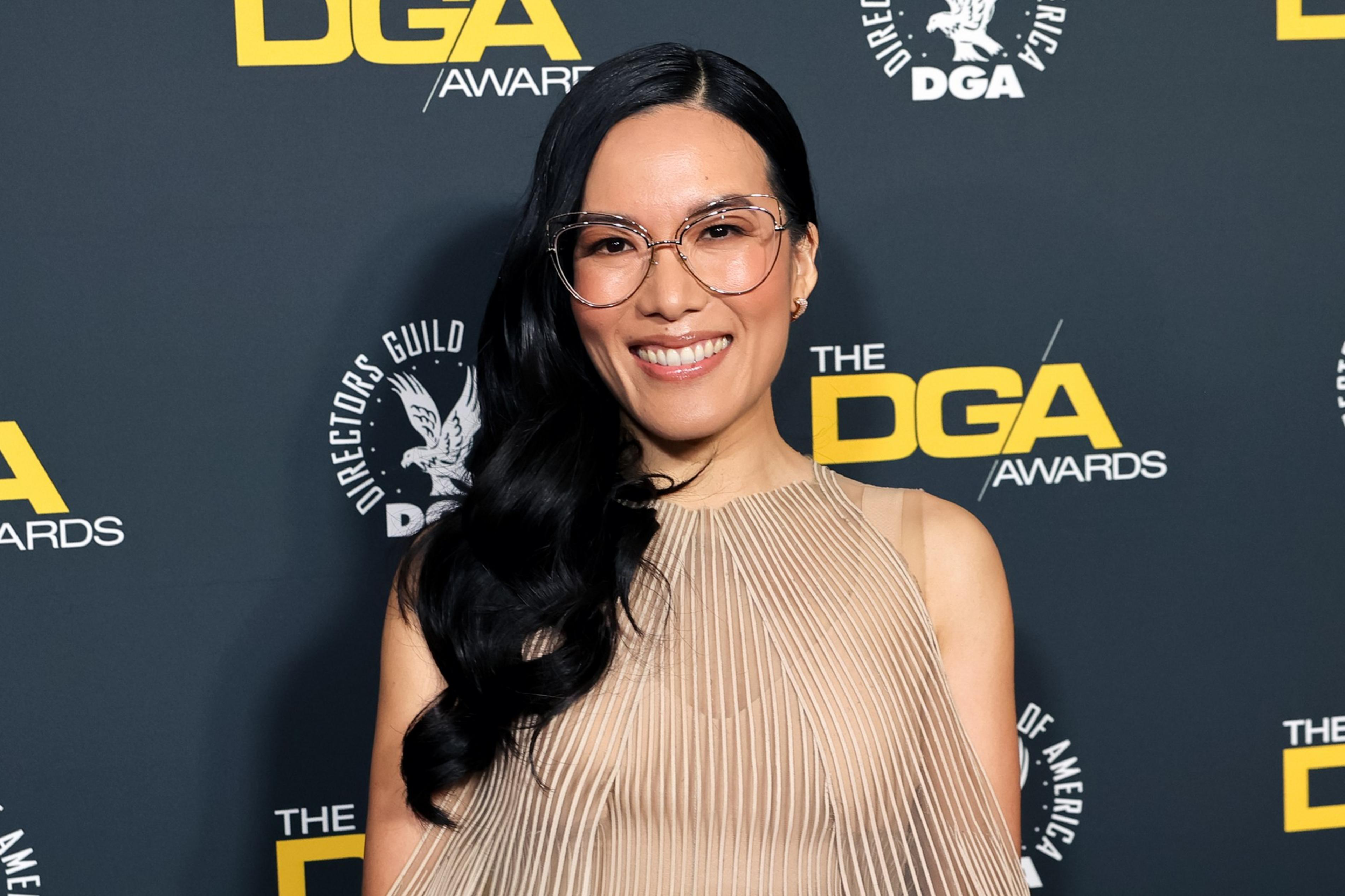 A person with long dark hair, wearing glasses and a beige dress, smiles against a backdrop featuring yellow "DGA Awards" text and logos.