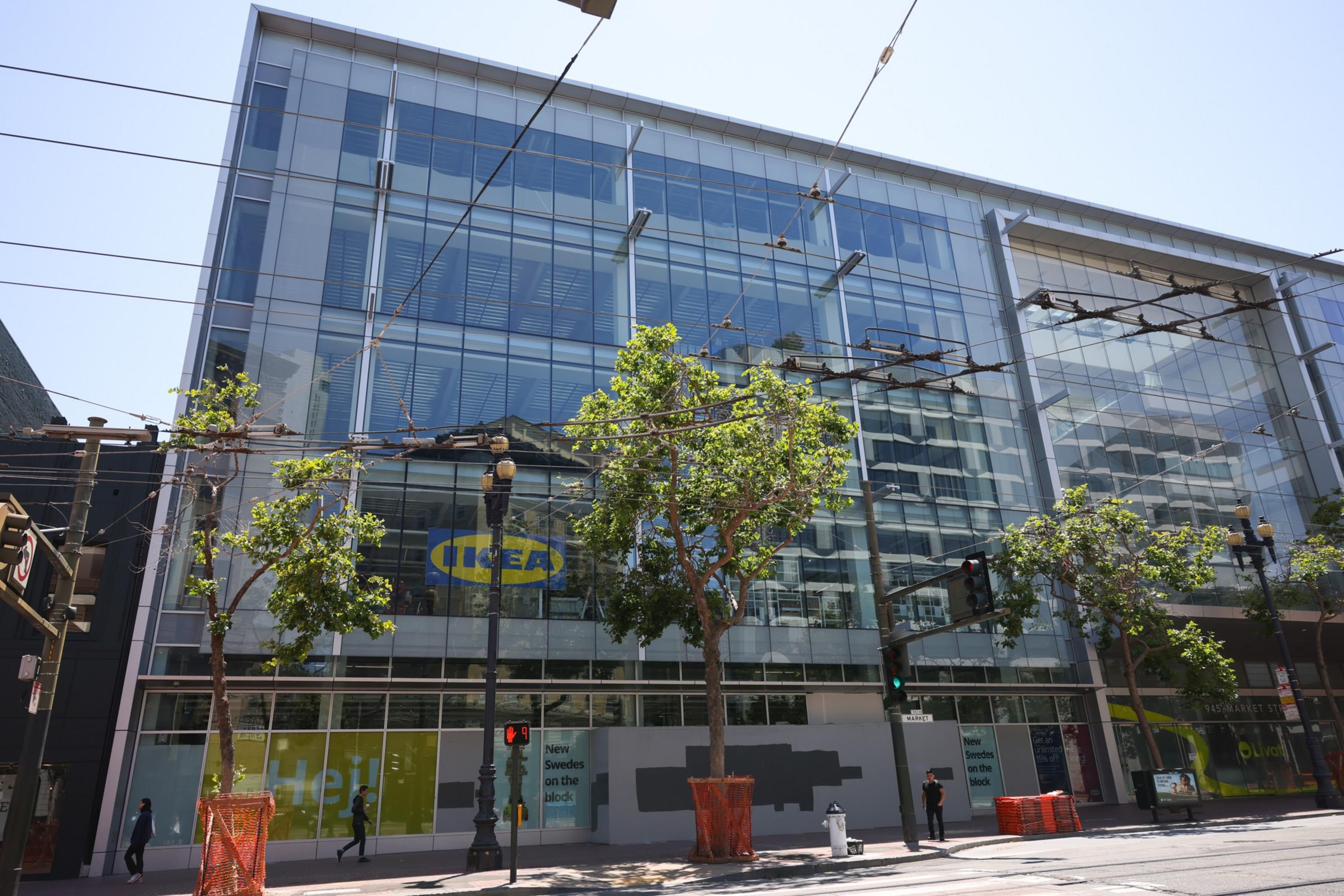 A modern glass-fronted IKEA store with trees planted along the sidewalk, streetcar wires overhead, and a few pedestrians walking nearby.