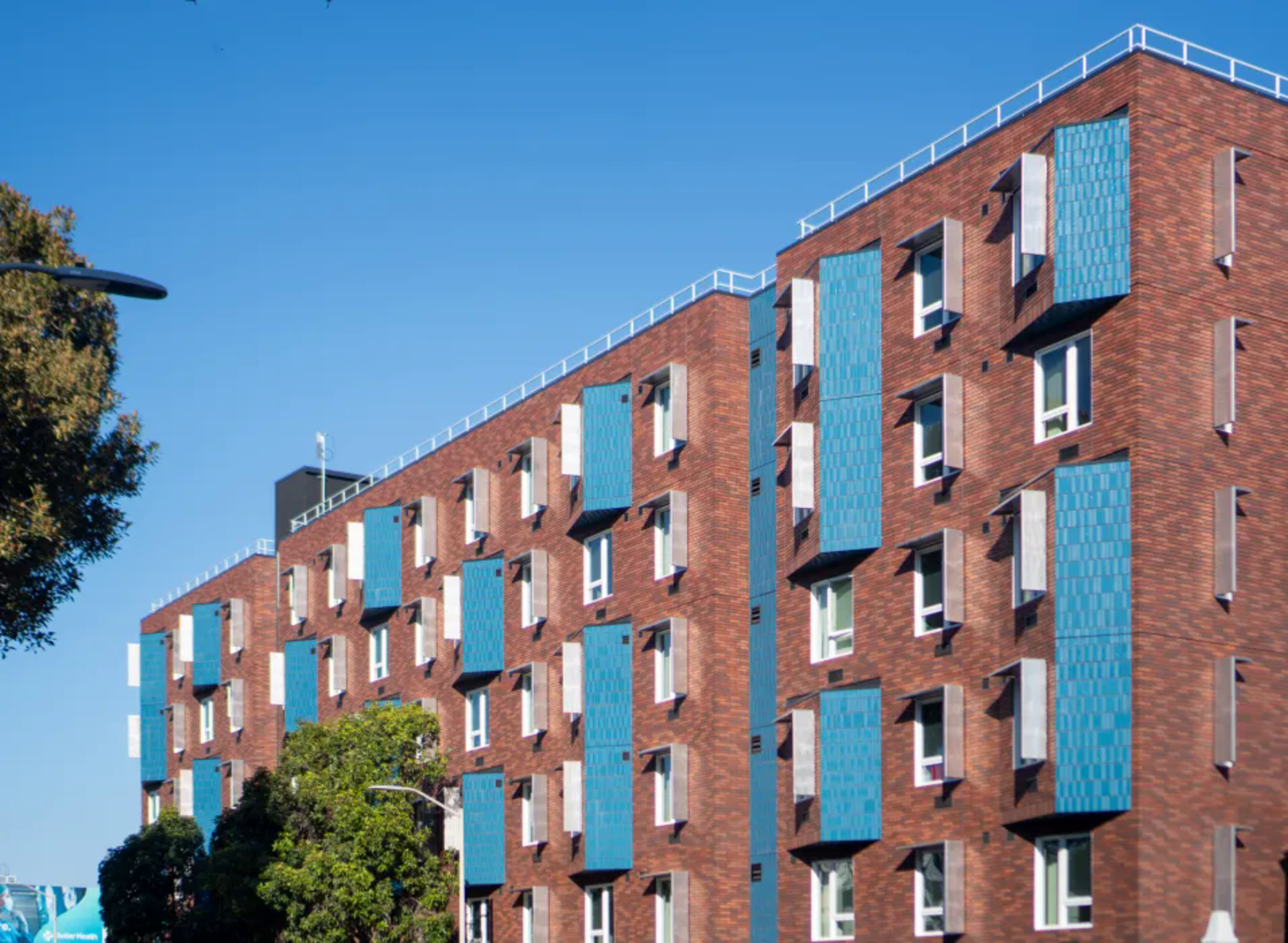 A red brick apartment building features multiple rows of windows, each accented with vertical blue panels under a clear blue sky.