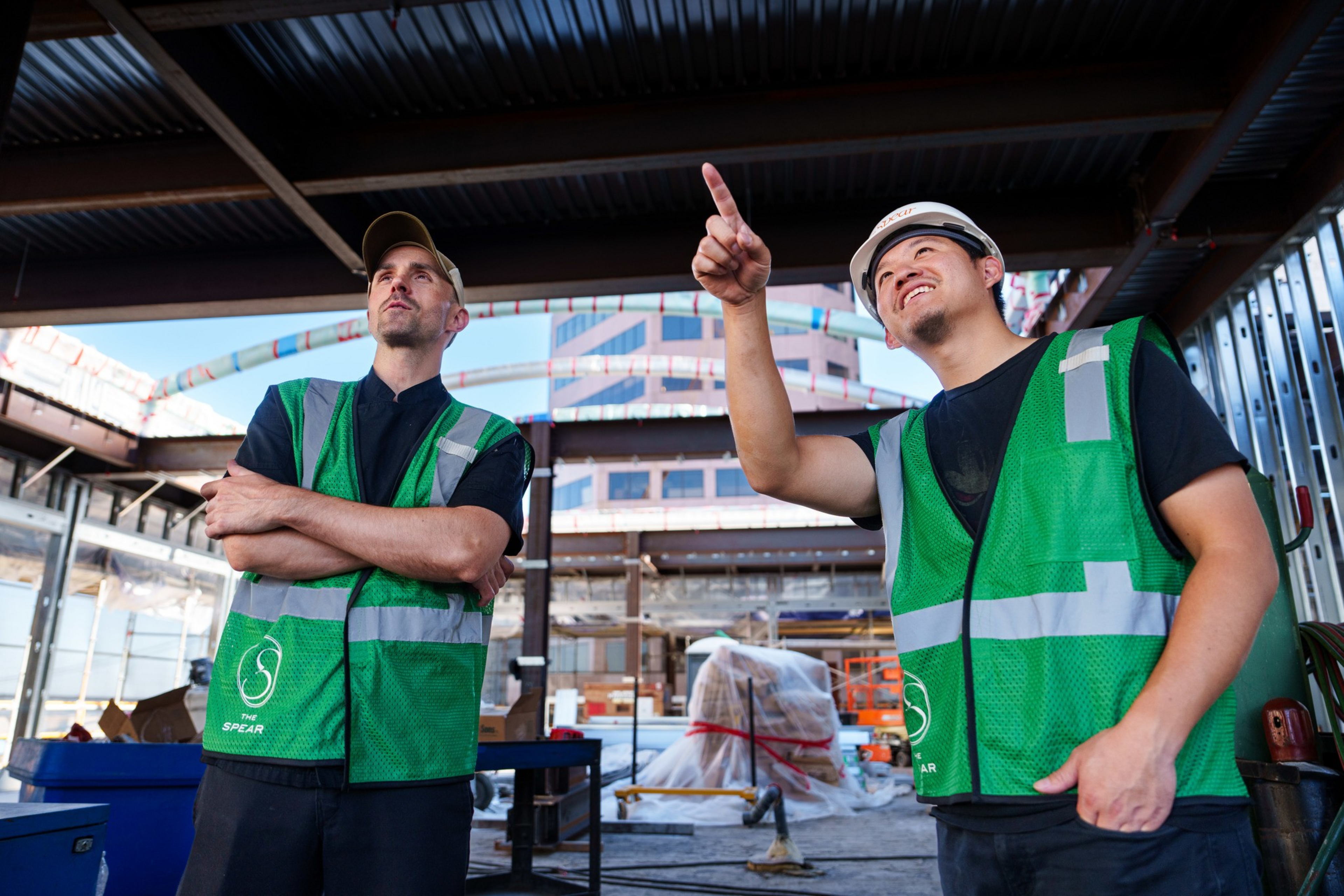 Two construction workers in green safety vests are on-site, one pointing upward while the other watches attentively with arms crossed.