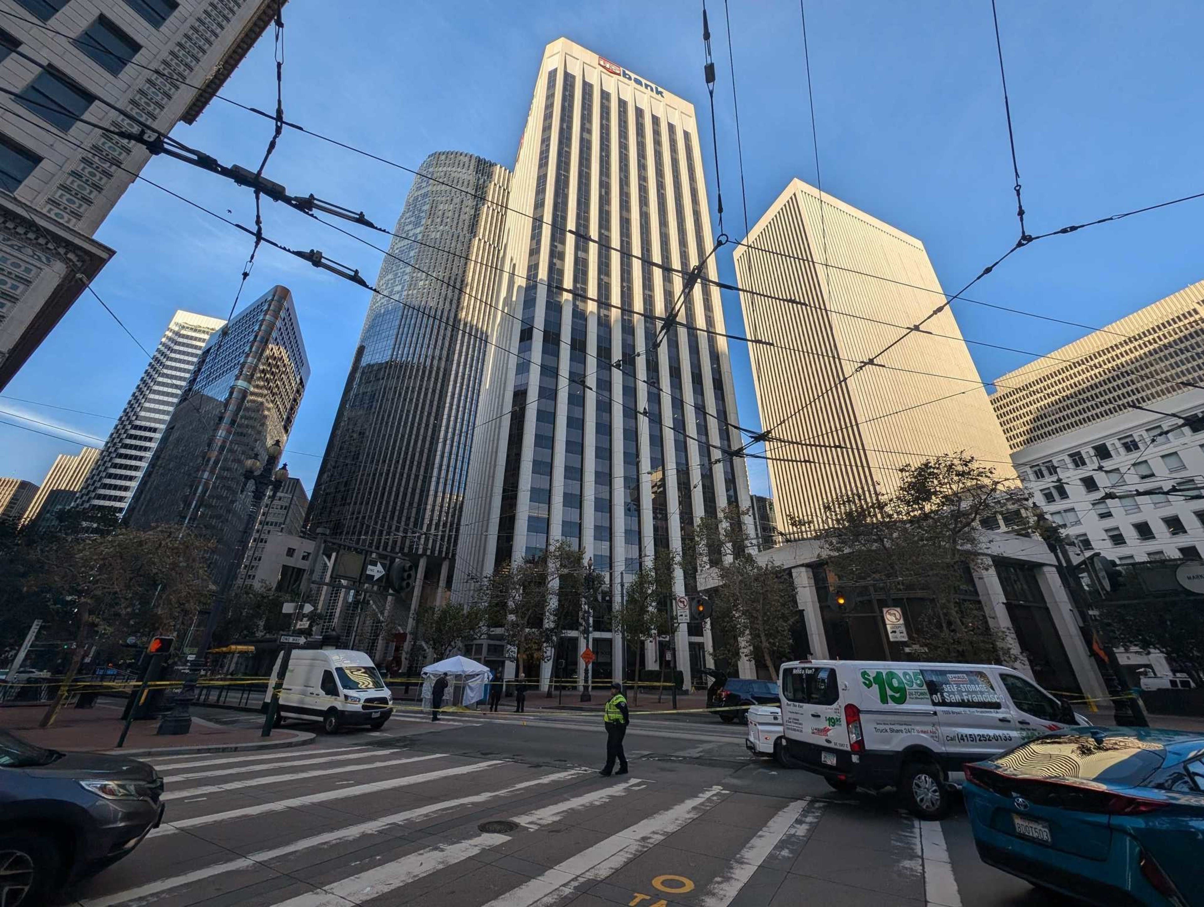 The image shows a city street with several tall skyscrapers. There are vehicles, a person directing traffic, and overhead power lines crossing the scene.