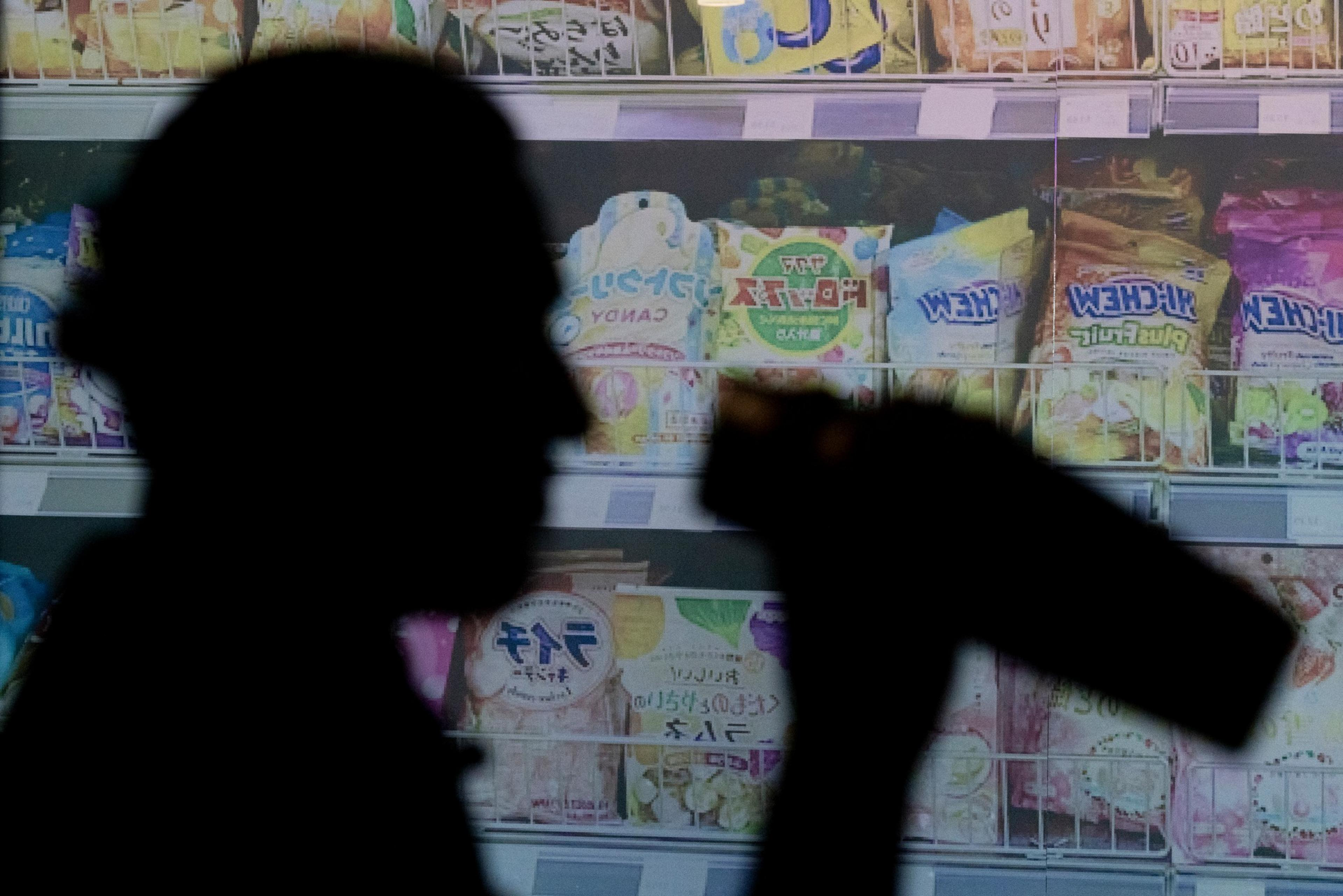 The image shows a silhouette of a person raising a bottle in front of a brightly lit store shelf filled with various colorful packaged products.