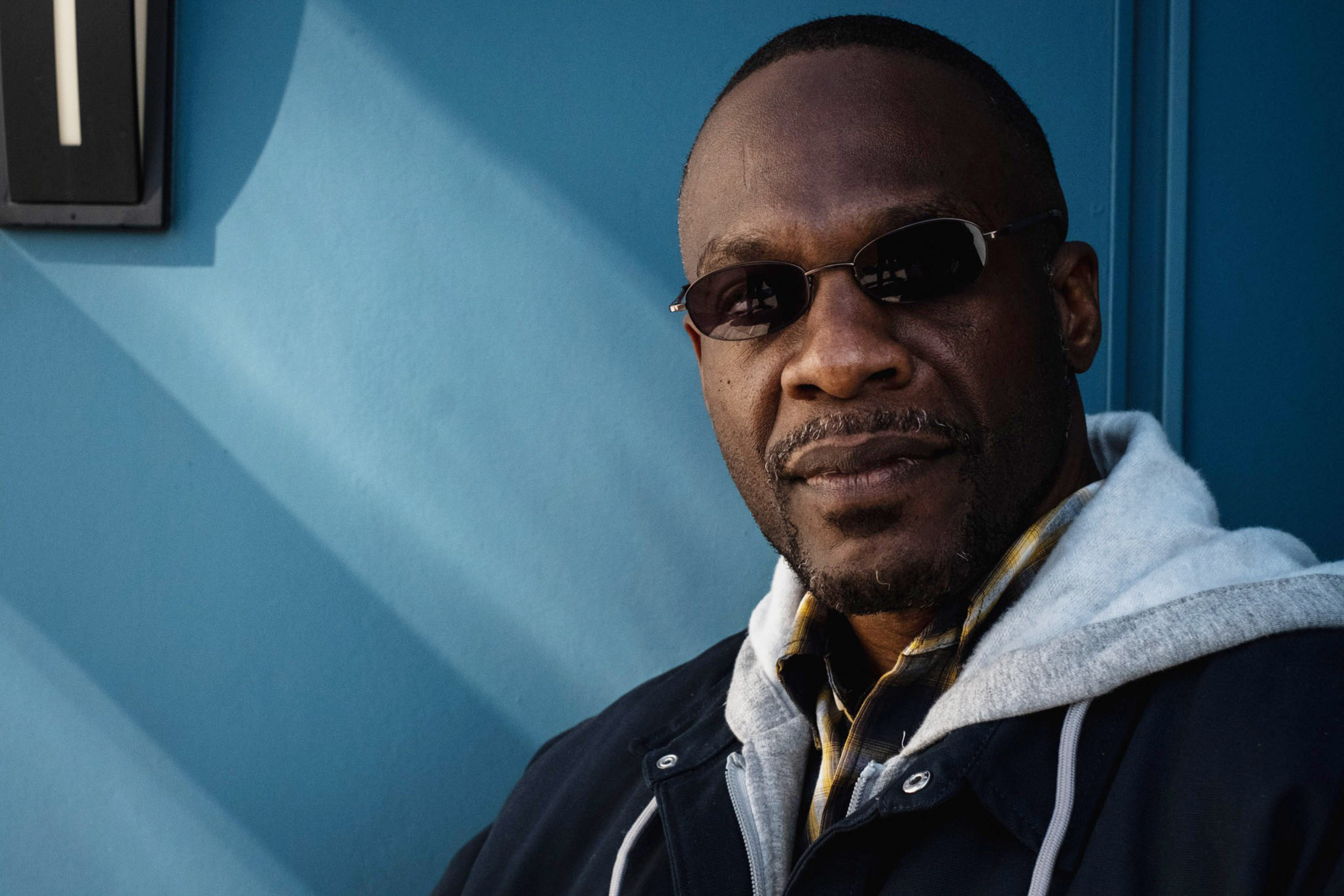 A man with dark sunglasses wears a gray hoodie layered under a dark jacket, standing against a blue wall with diagonal shadow lines.