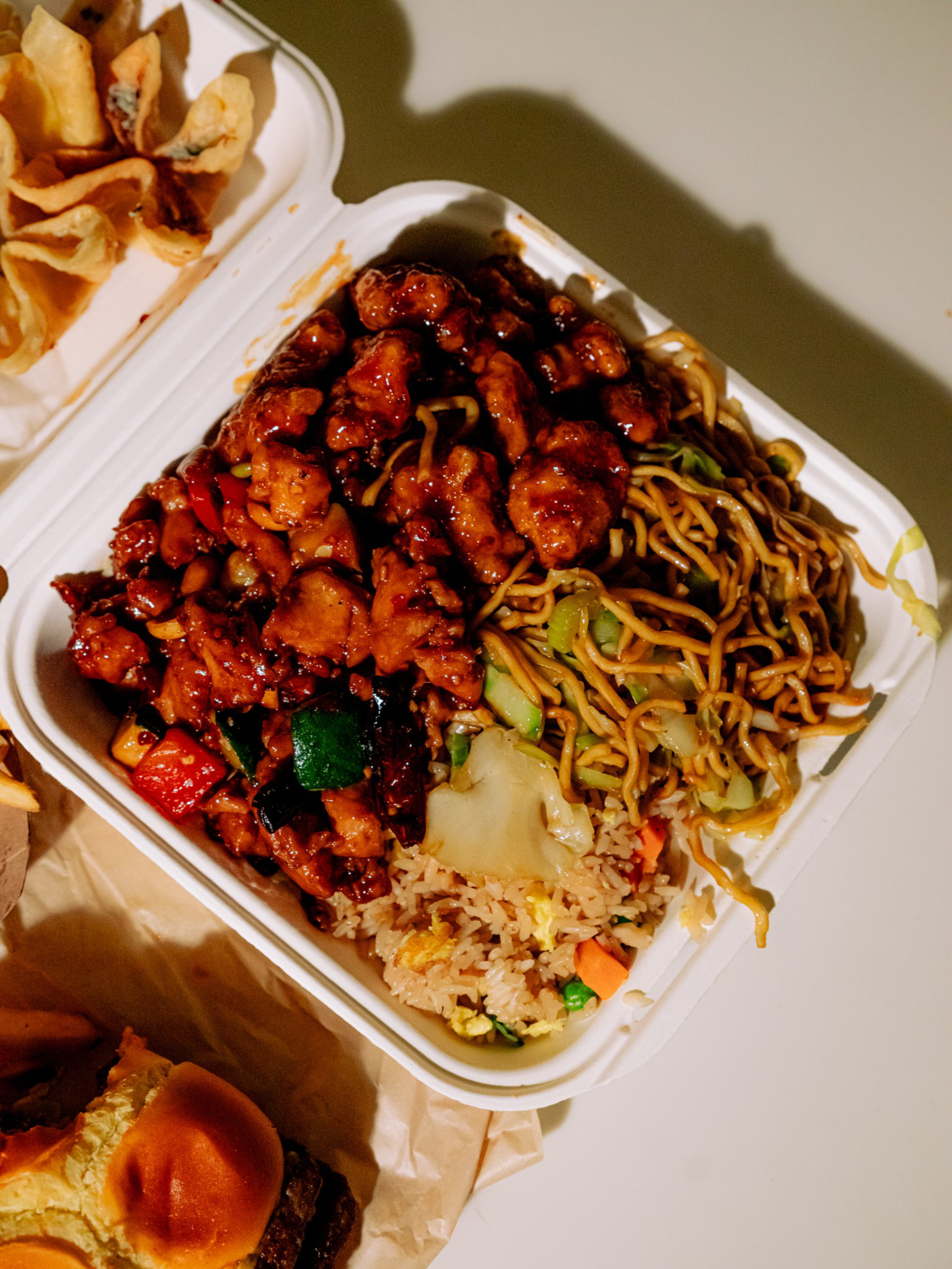Sweet and sour chicken, stir-fried noodles with vegetables, and fried rice with peas and carrots in a white takeout container, next to fried wontons and a sandwich.