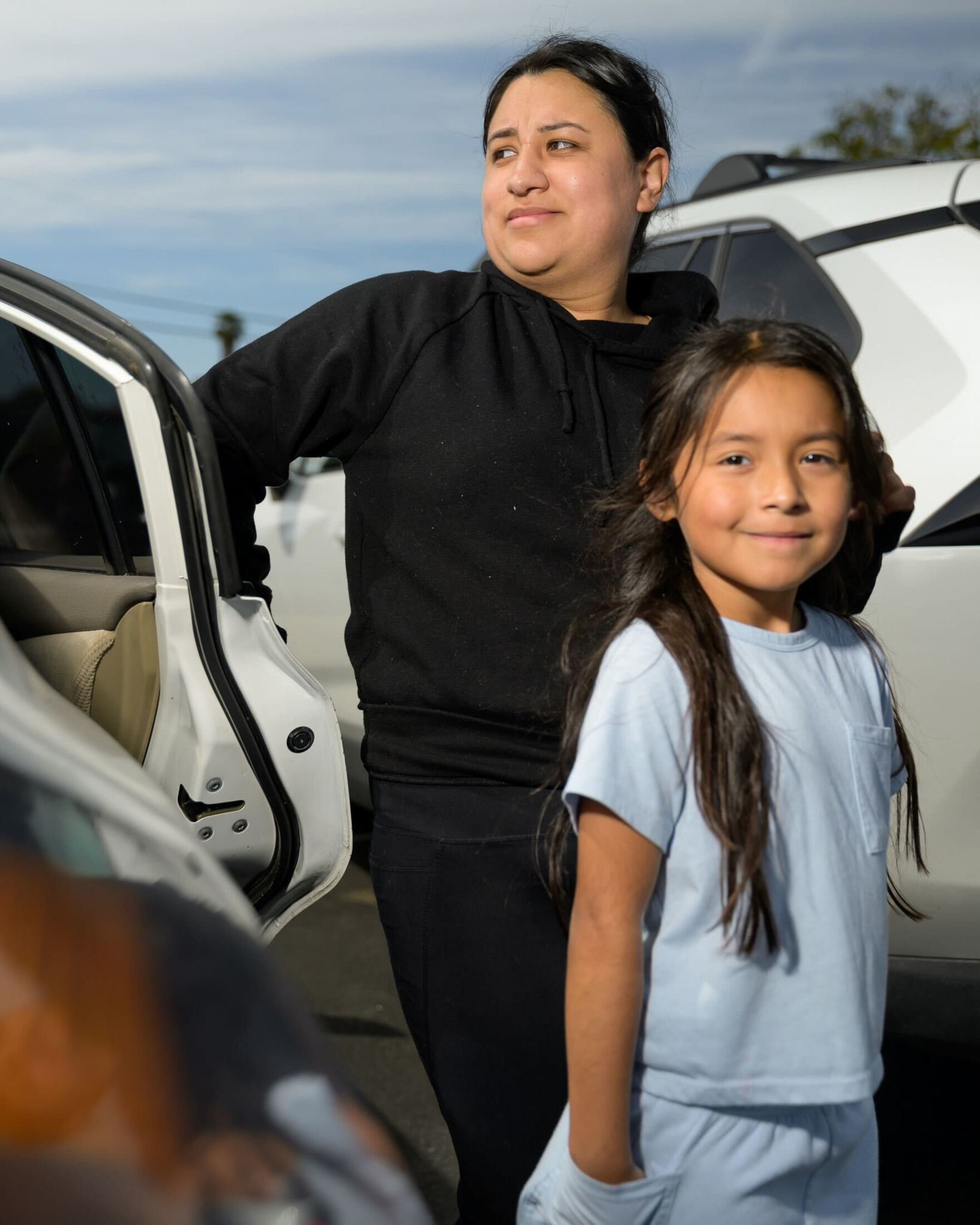 A woman in a black hoodie stands near an open car door, while a young girl in a light blue outfit smiles in front of her on a sunny day.