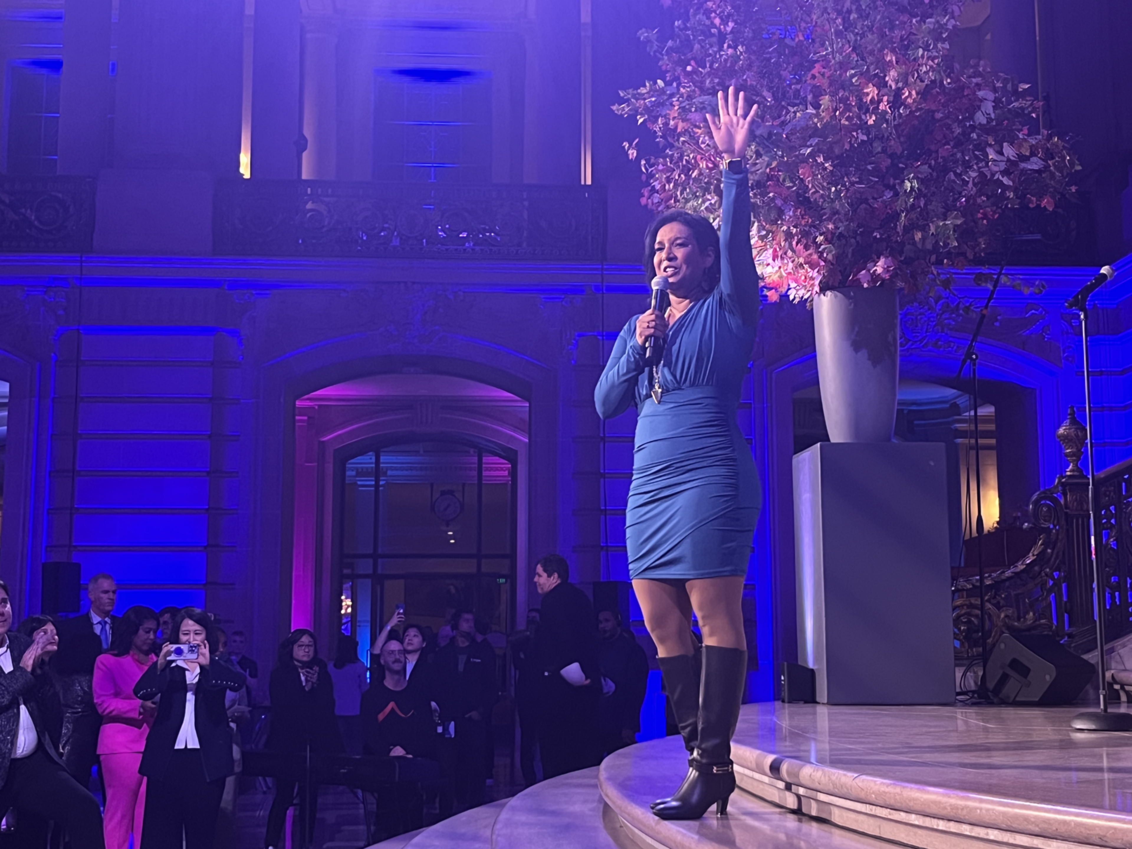A woman in a blue dress and boots speaks using a microphone