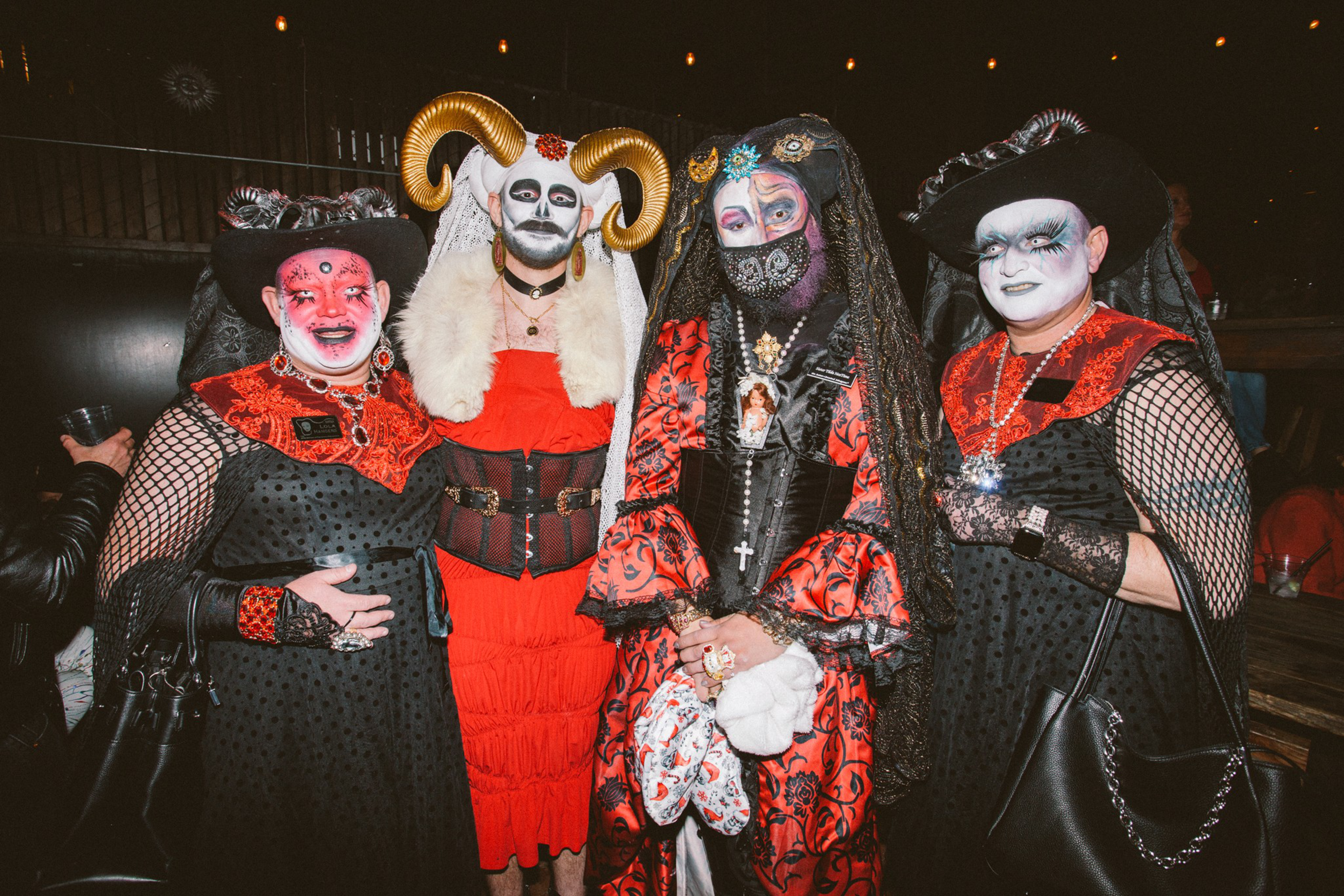 Four people dressed in elaborate black and red costumes with dramatic makeup and accessories, including horns, veils, and hats, pose indoors at night.