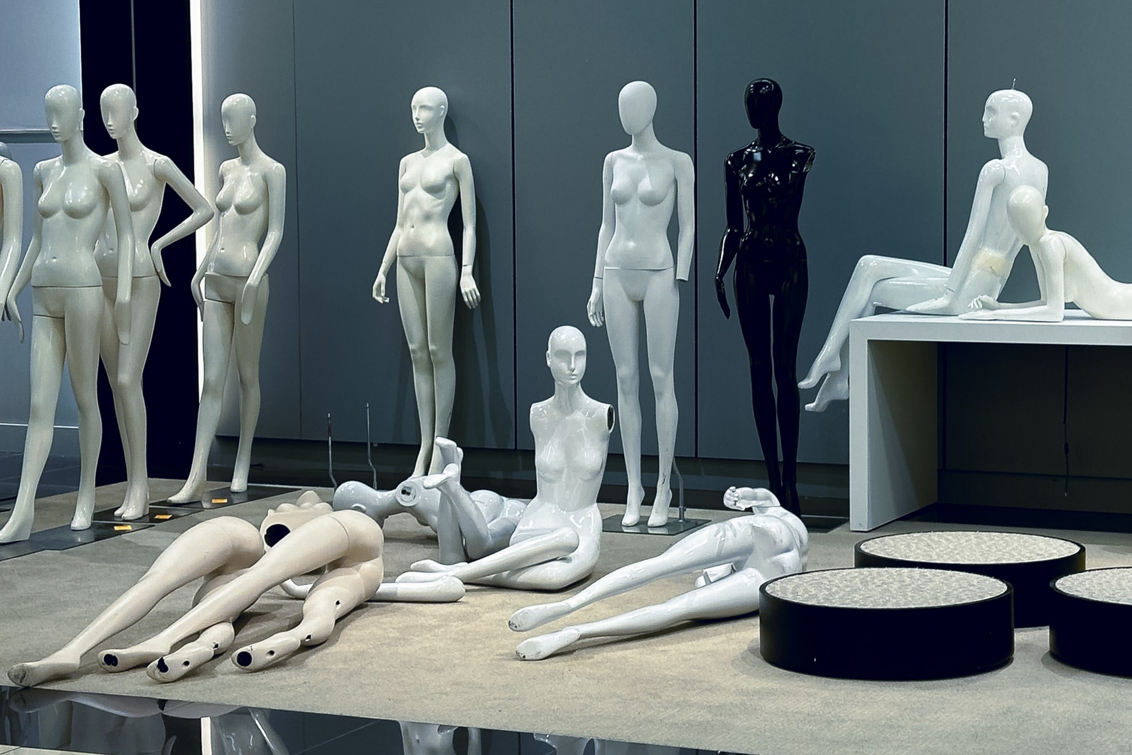 The image shows several white and one black mannequin, some standing and others disassembled or sitting on a carpeted floor in front of a gray wall.