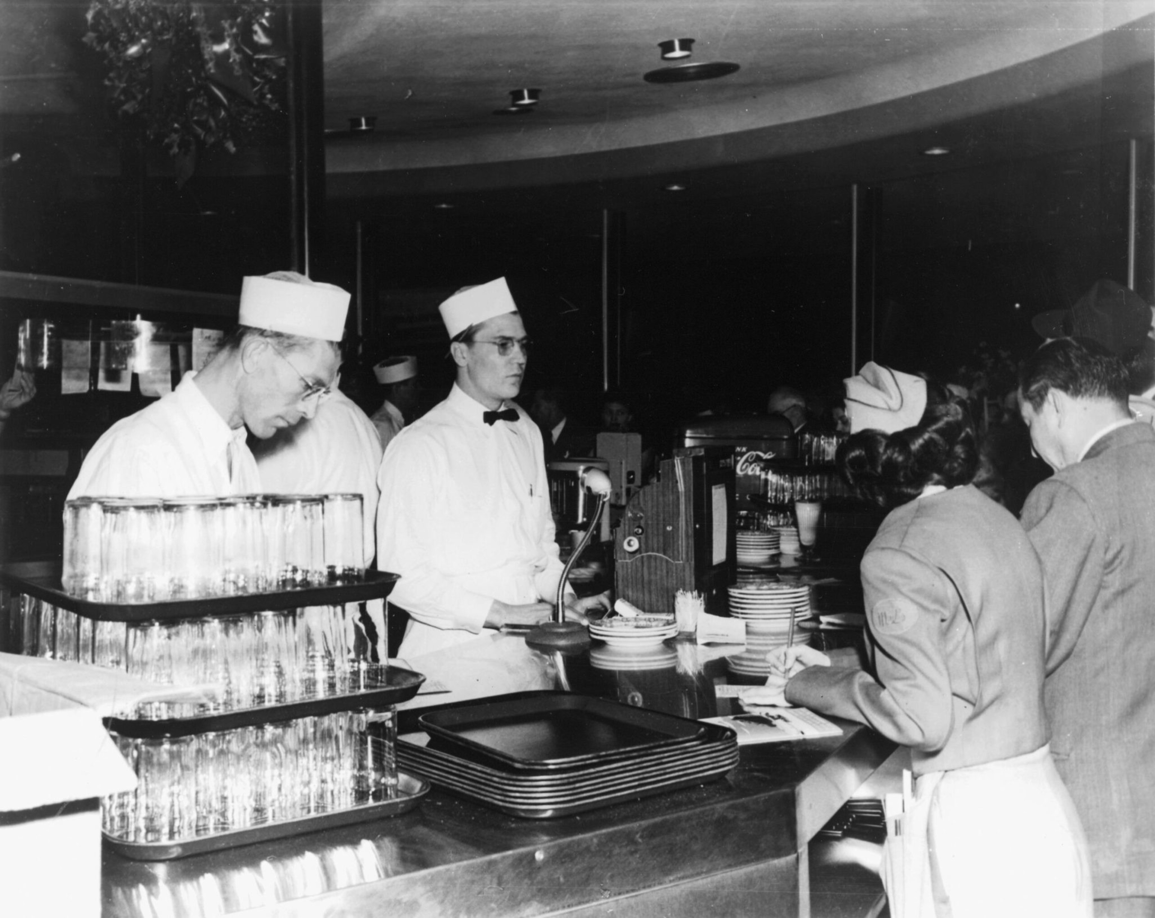 Two male servers in white uniforms and hats stand behind a diner counter, while a woman in a hat and a man wait on the customer side.