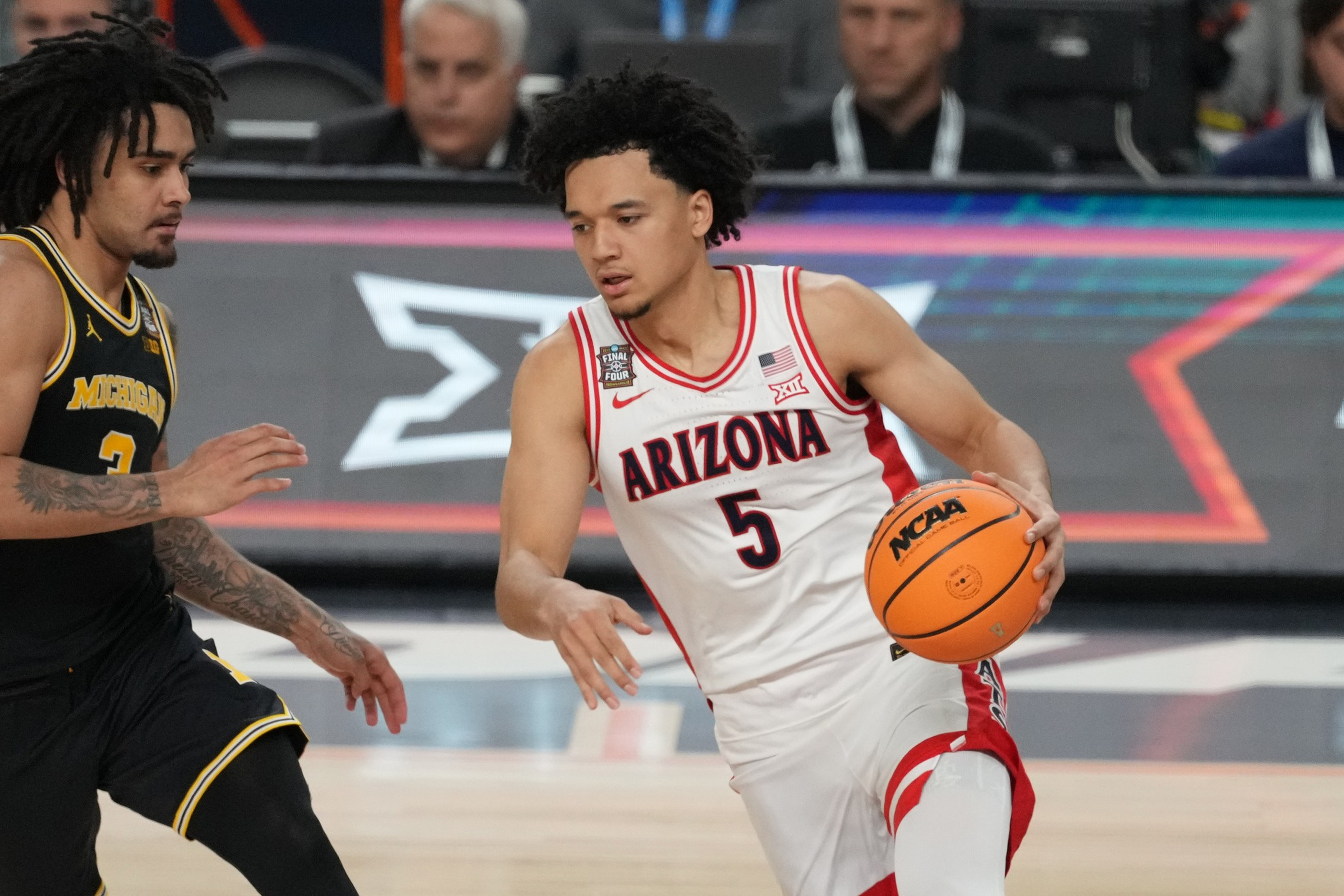 A basketball player wearing a white Arizona jersey dribbles the ball as a Michigan player in a black jersey defends him.
