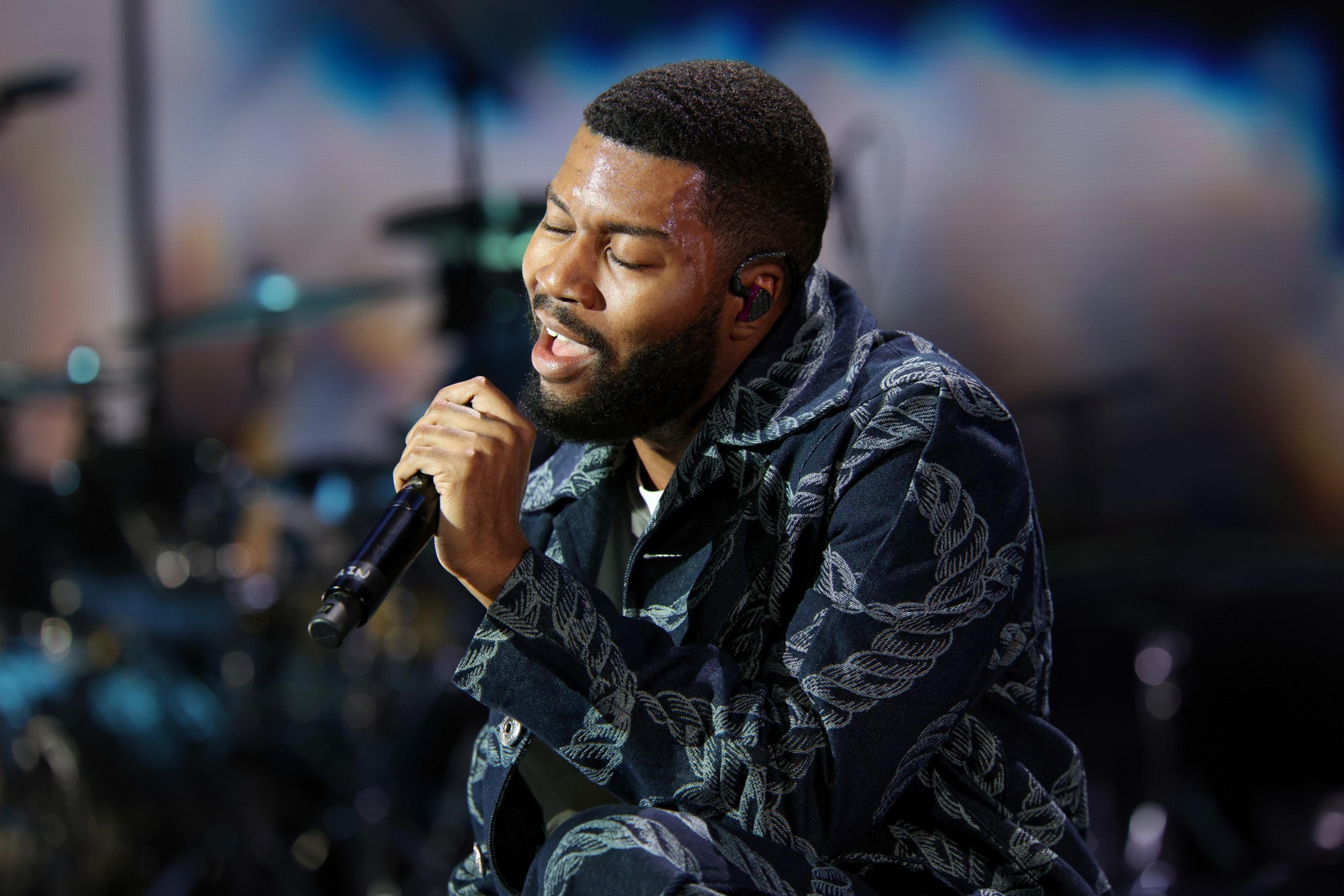 A bearded man with closed eyes passionately sings into a microphone while wearing a patterned jacket and ear monitors on stage.