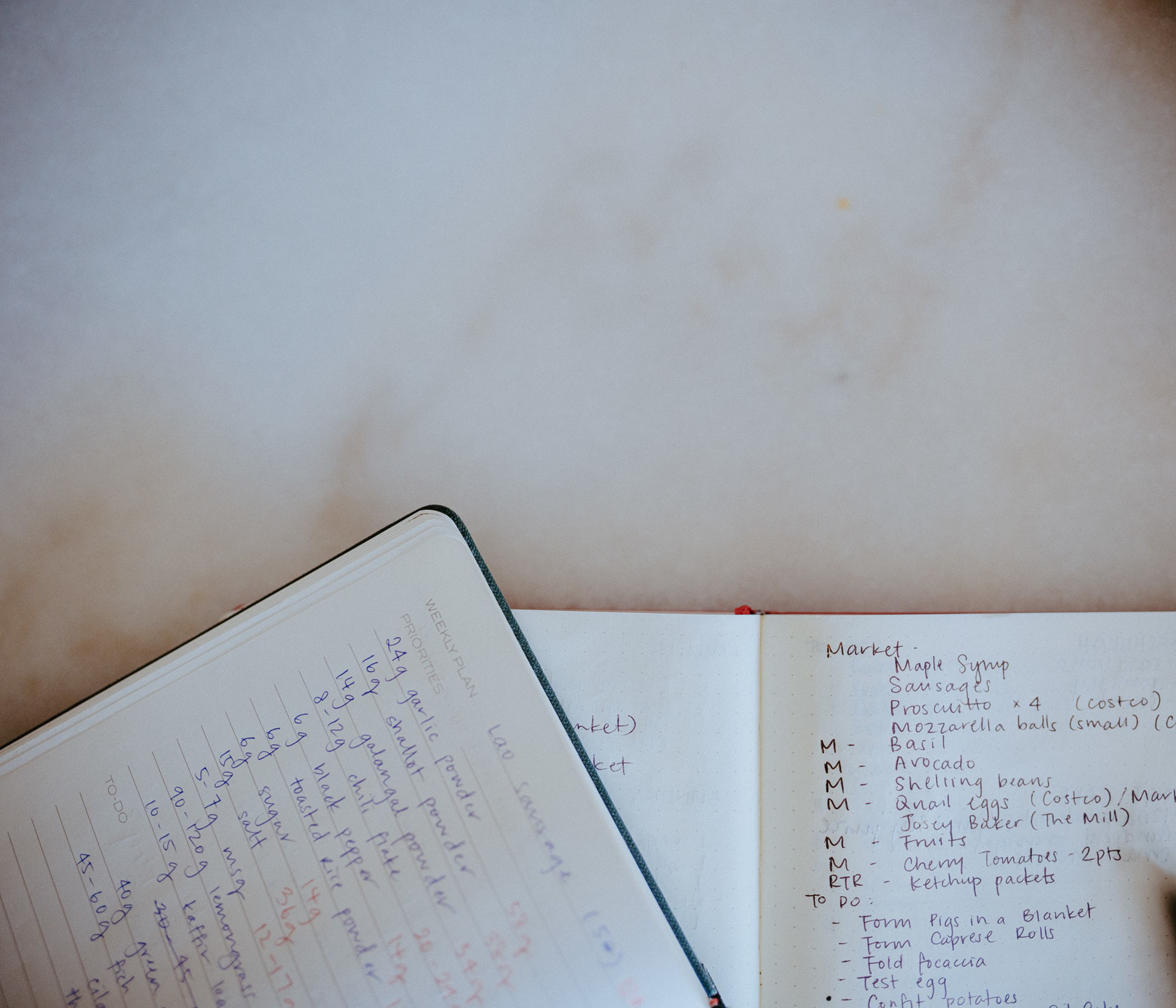 Two open notebooks lie on a marble surface, one showing a weekly plan with numbered tasks, the other containing a detailed grocery list and to-do notes.