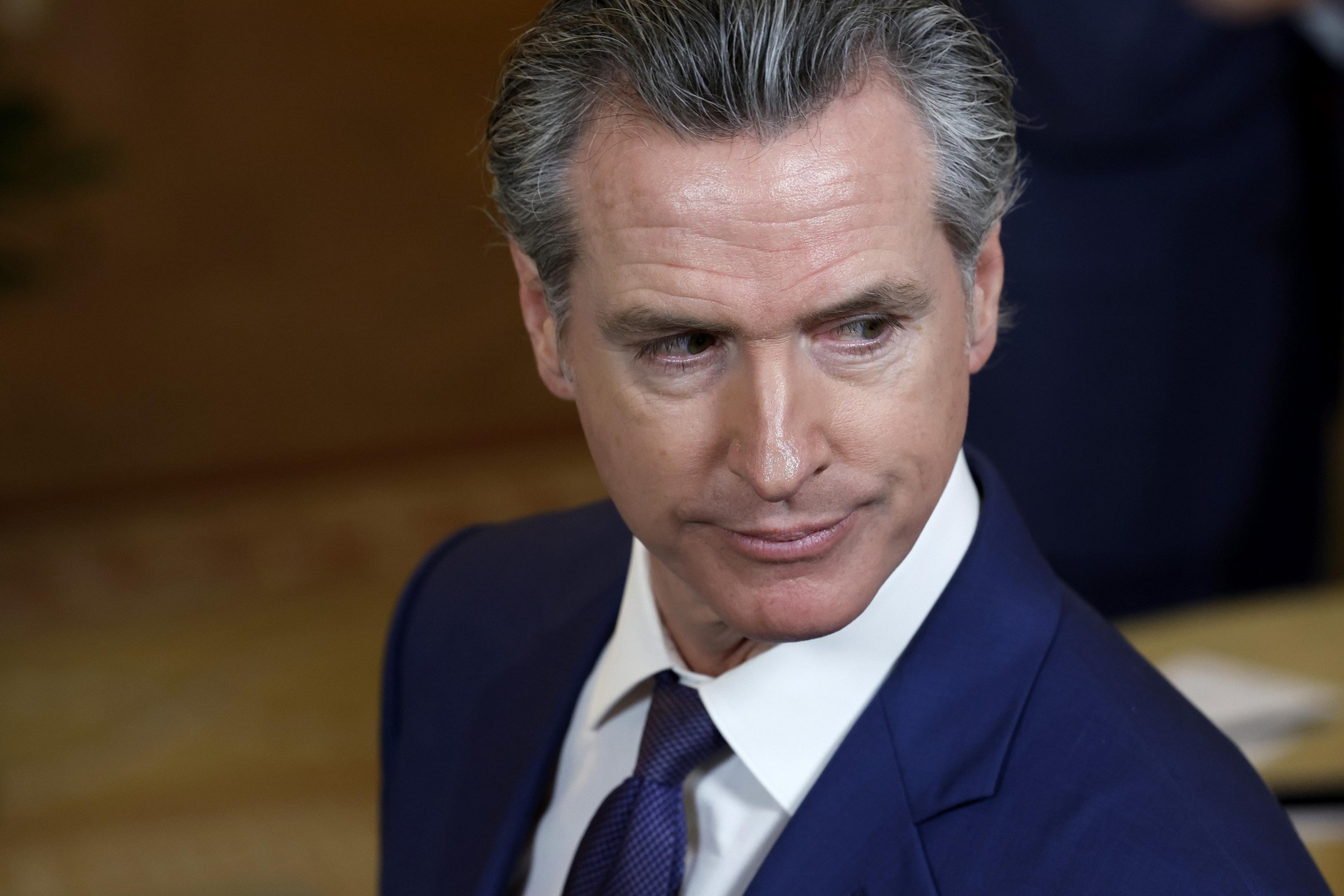 A man in a blue suit and tie looks to his left with a serious expression, set against a blurred, warm-toned background.