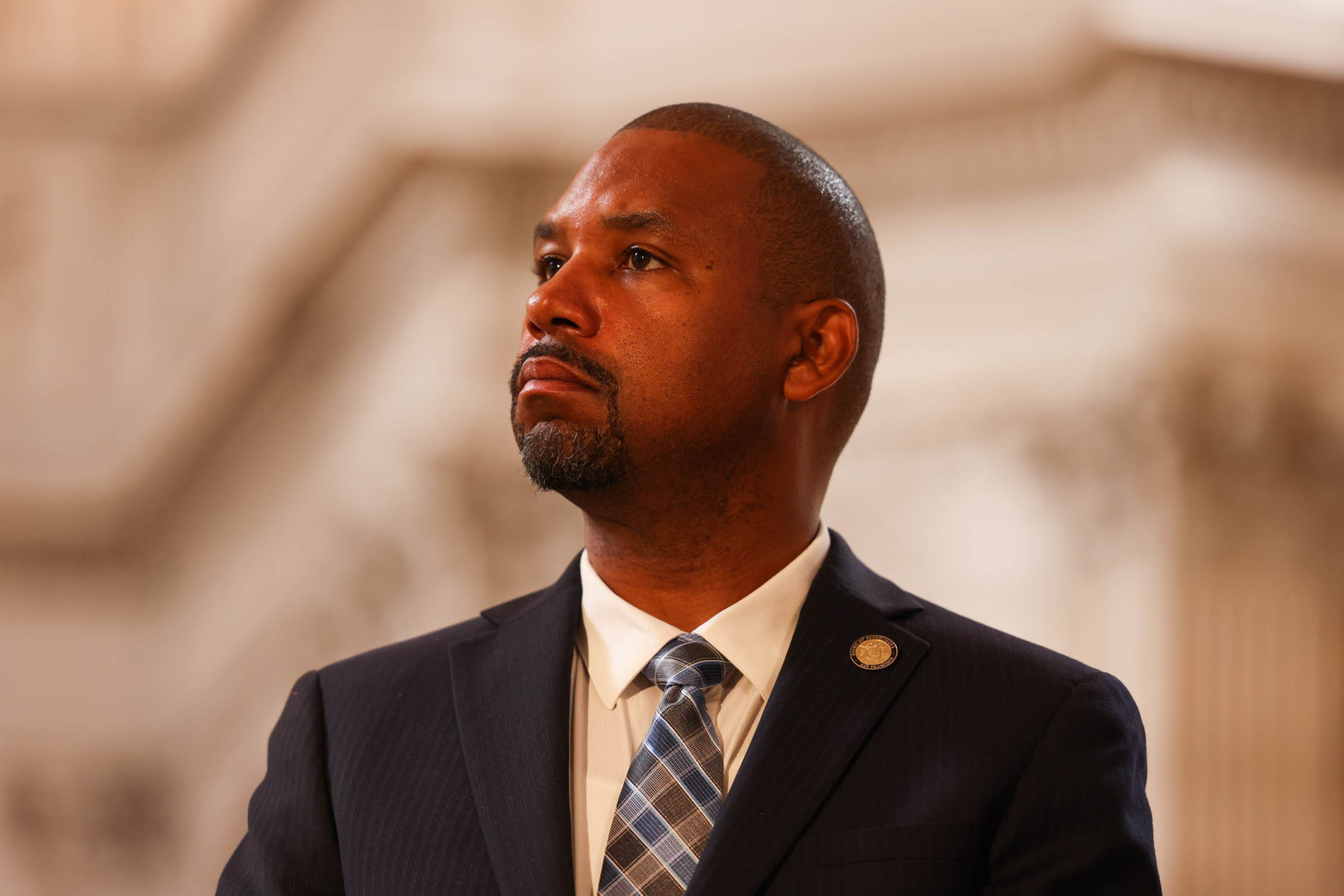 A man in a suit with a pin on his lapel stands in a formal setting. He looks upward with a serious expression, and he wears a checkered tie.