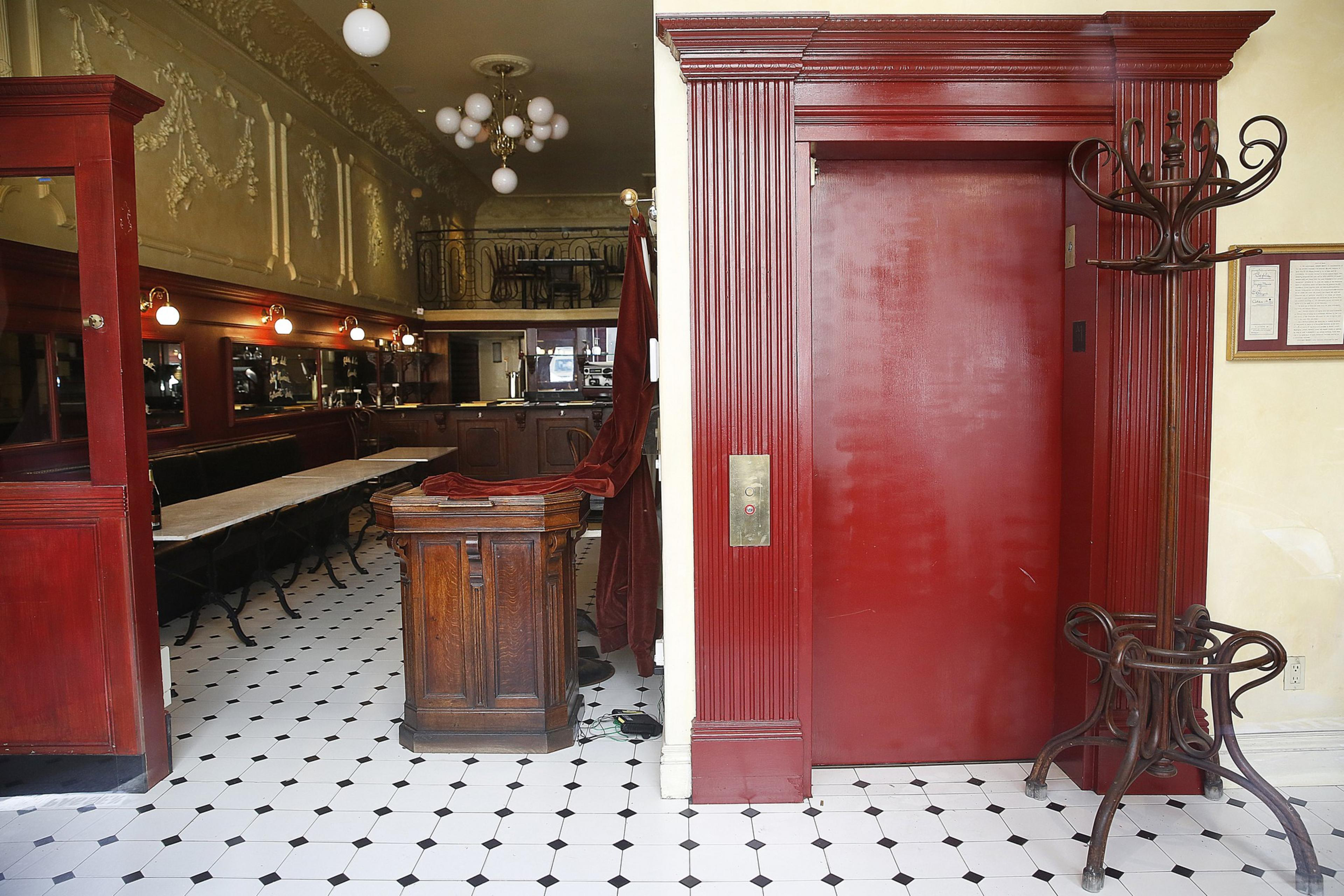A red elevator door with a vintage wooden coat rack stands next to a dining area with long tables, chairs, chandeliers, and decorative molding.