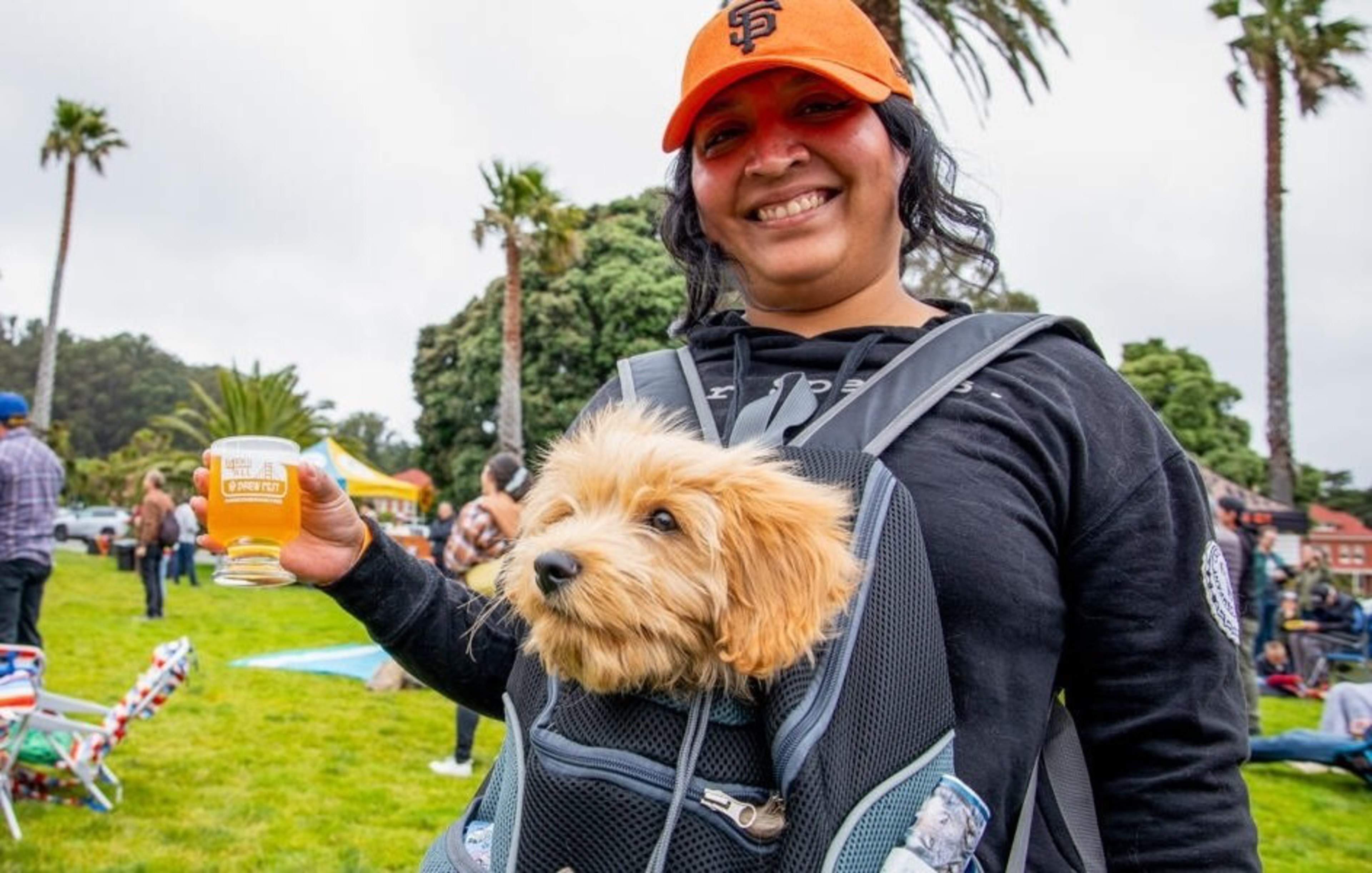 A smiling woman wearing an orange hat holds a glass of beer while carrying a small fluffy dog in a front-facing backpack at an outdoor event.
