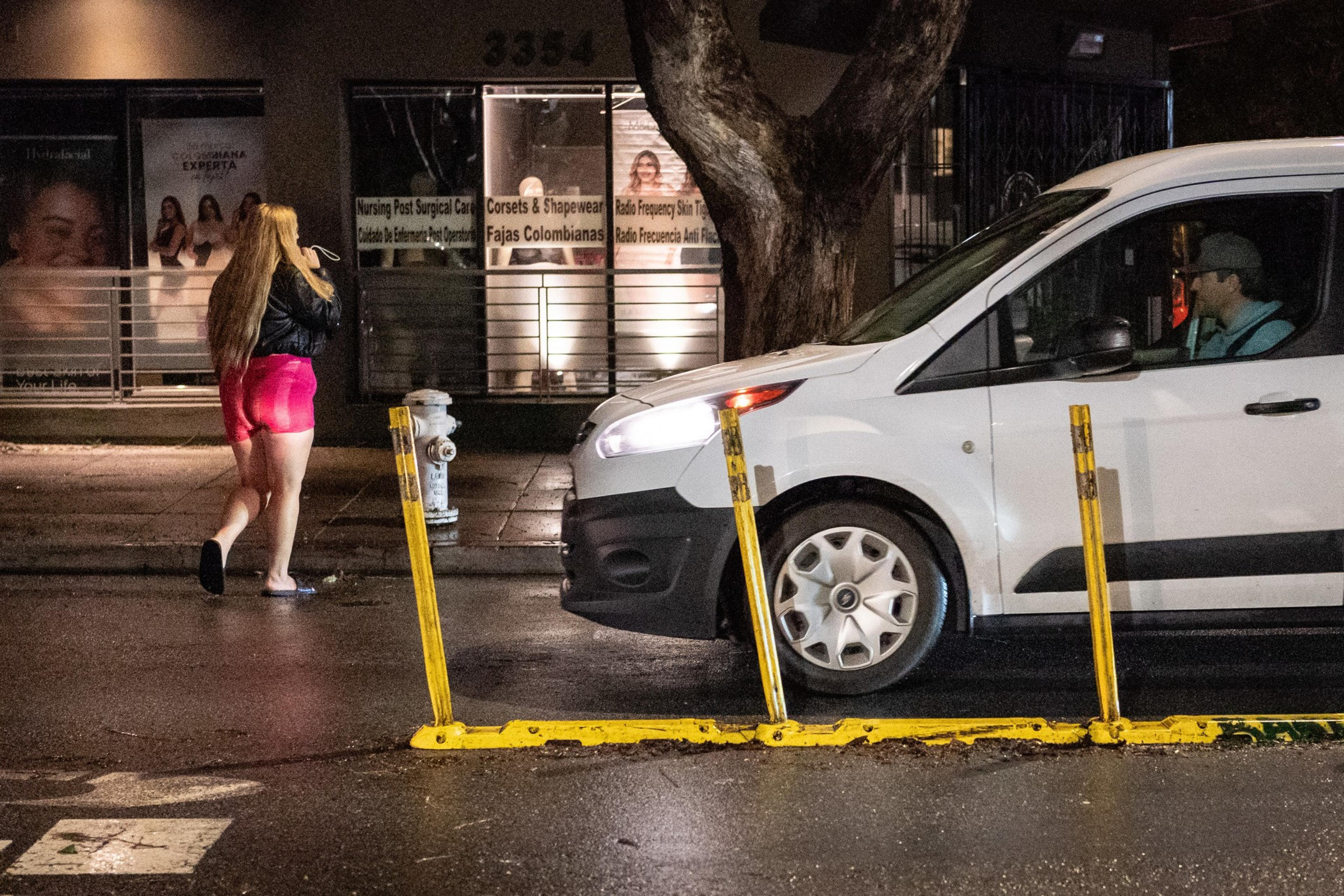 A person with long hair, a dark top and pink shorts crosses a rain-soaked street at nighttime in front of a stopped white car.