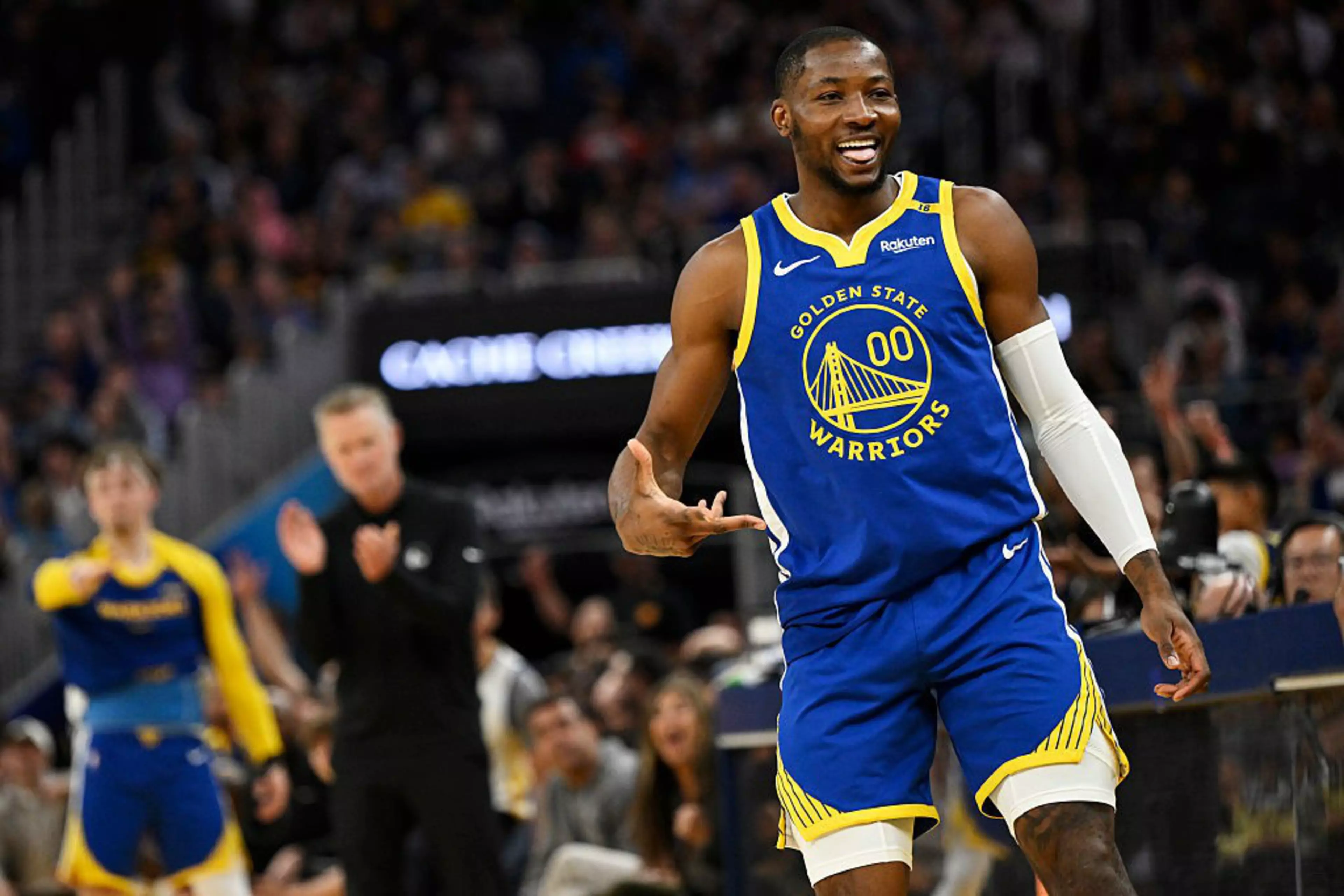 A basketball player in a Golden State Warriors uniform smiles on the court. The crowd is blurred in the background, and others on the sidelines cheer him on.