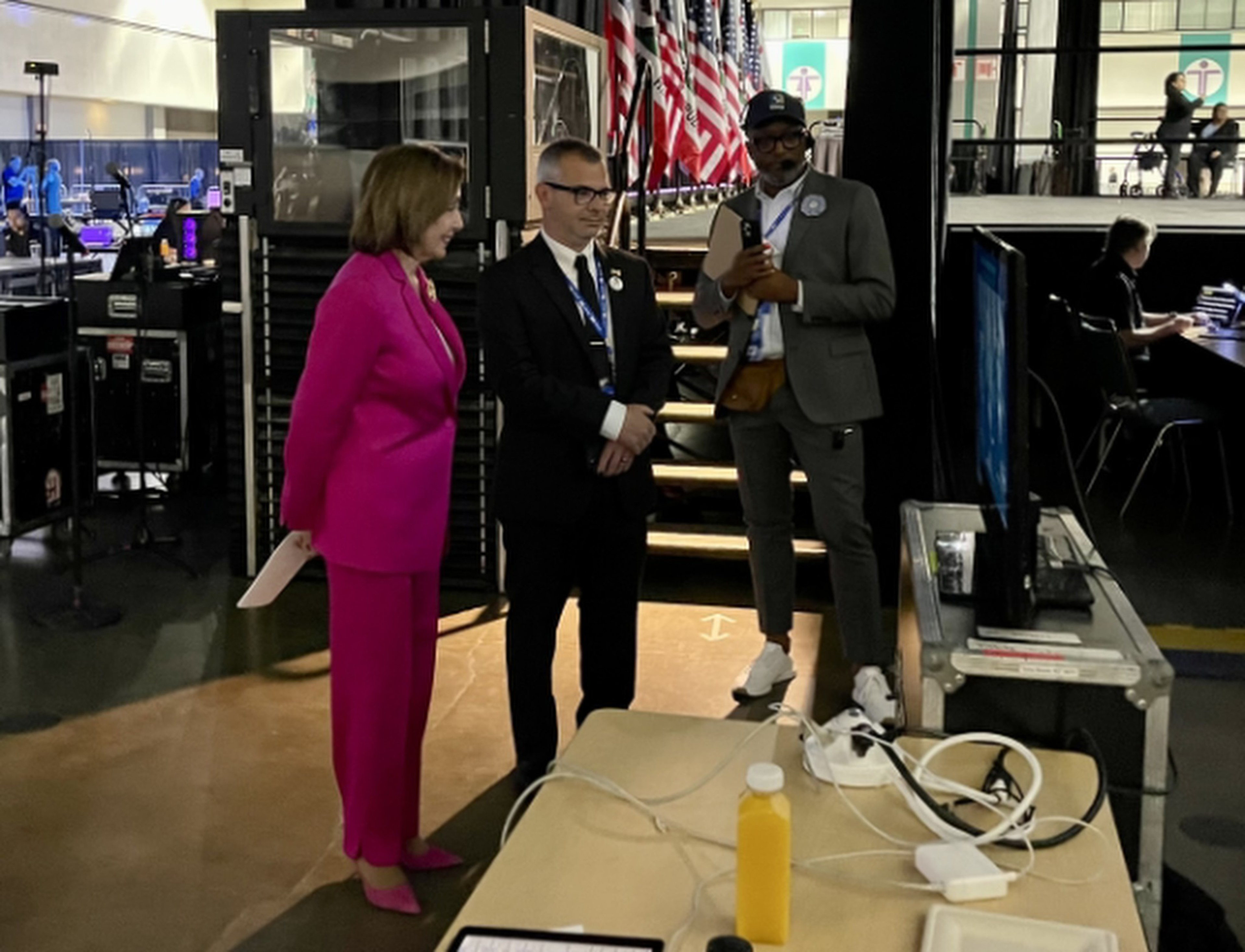 Three individuals stand backstage near a table with wires and an orange drink, with a row of American flags and people seated at tables in the background.
