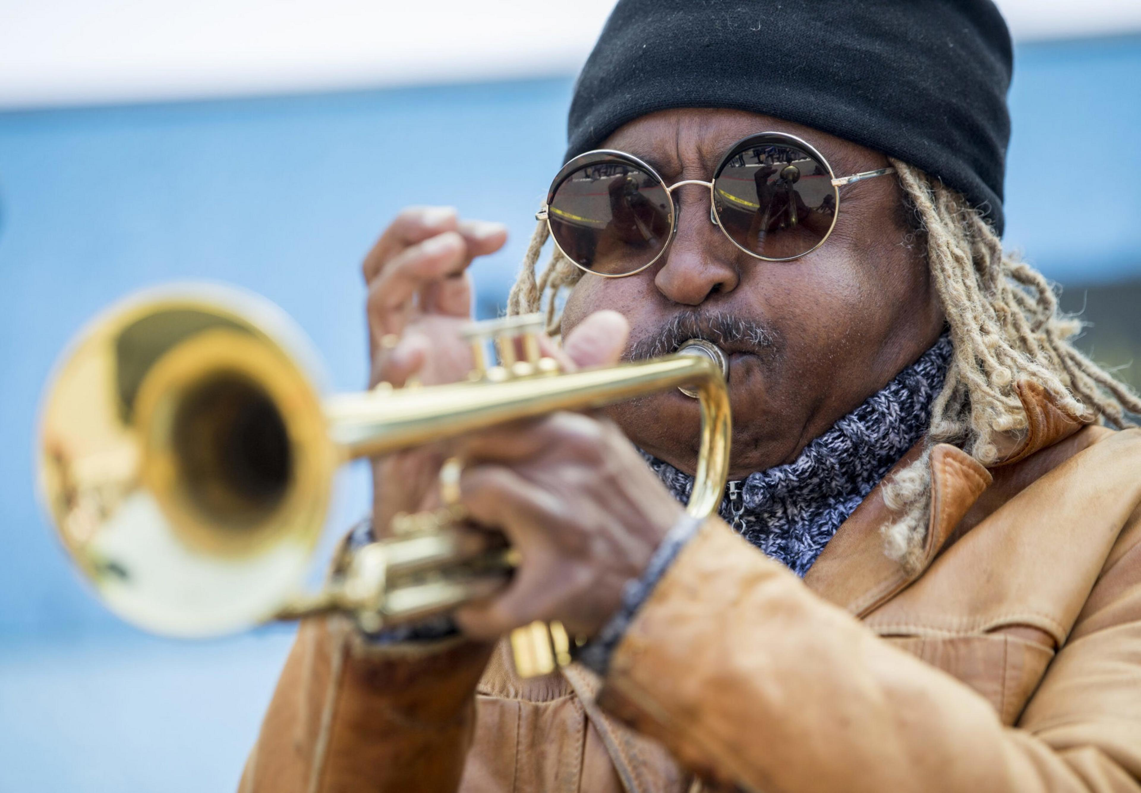 A person with sunglasses and a black beanie plays a trumpet passionately. They are wearing a tan coat and have light-colored braided hair.