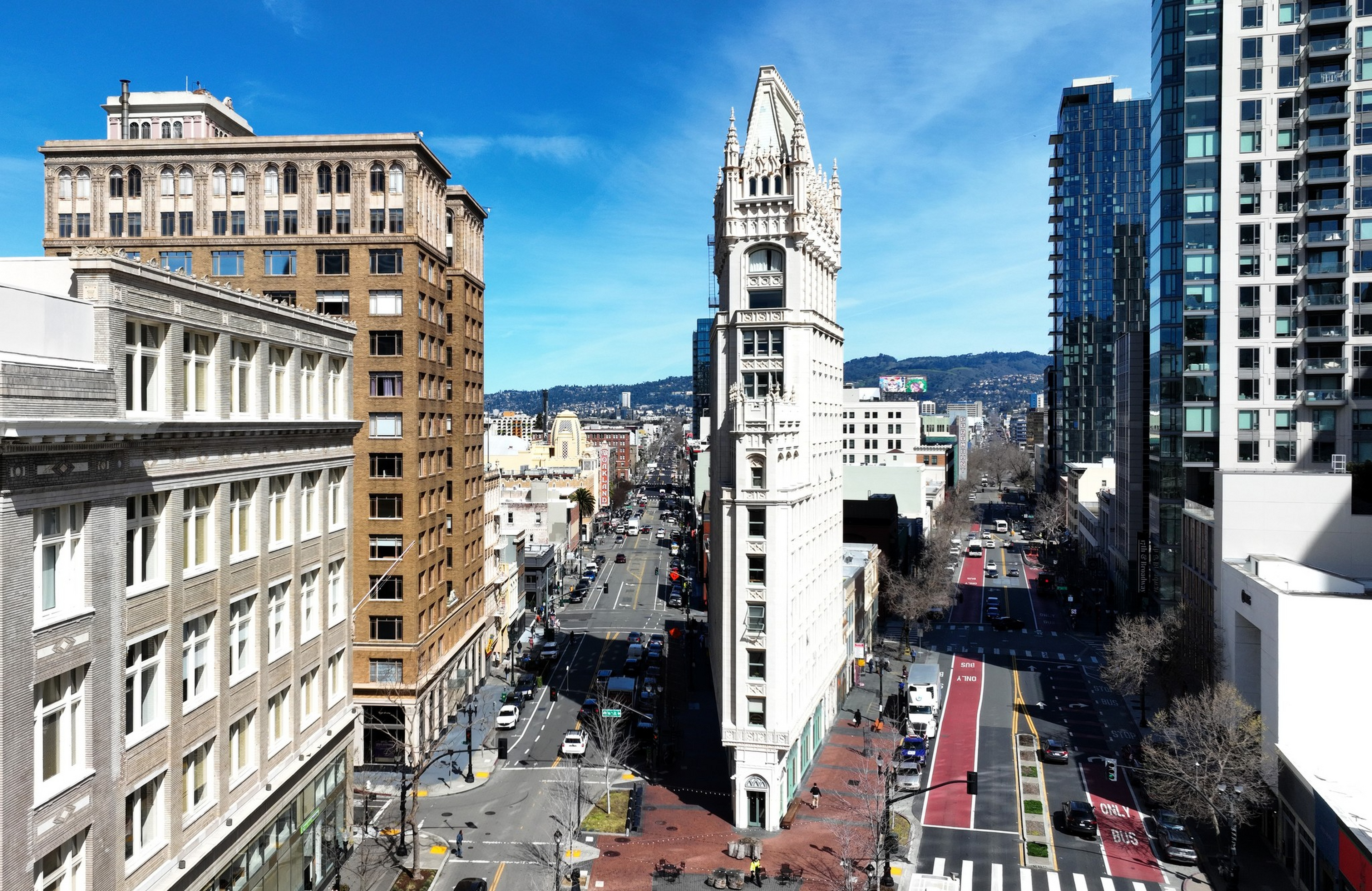 A narrow, pointed white clock tower stands at a busy intersection between tall buildings under a clear blue sky.
