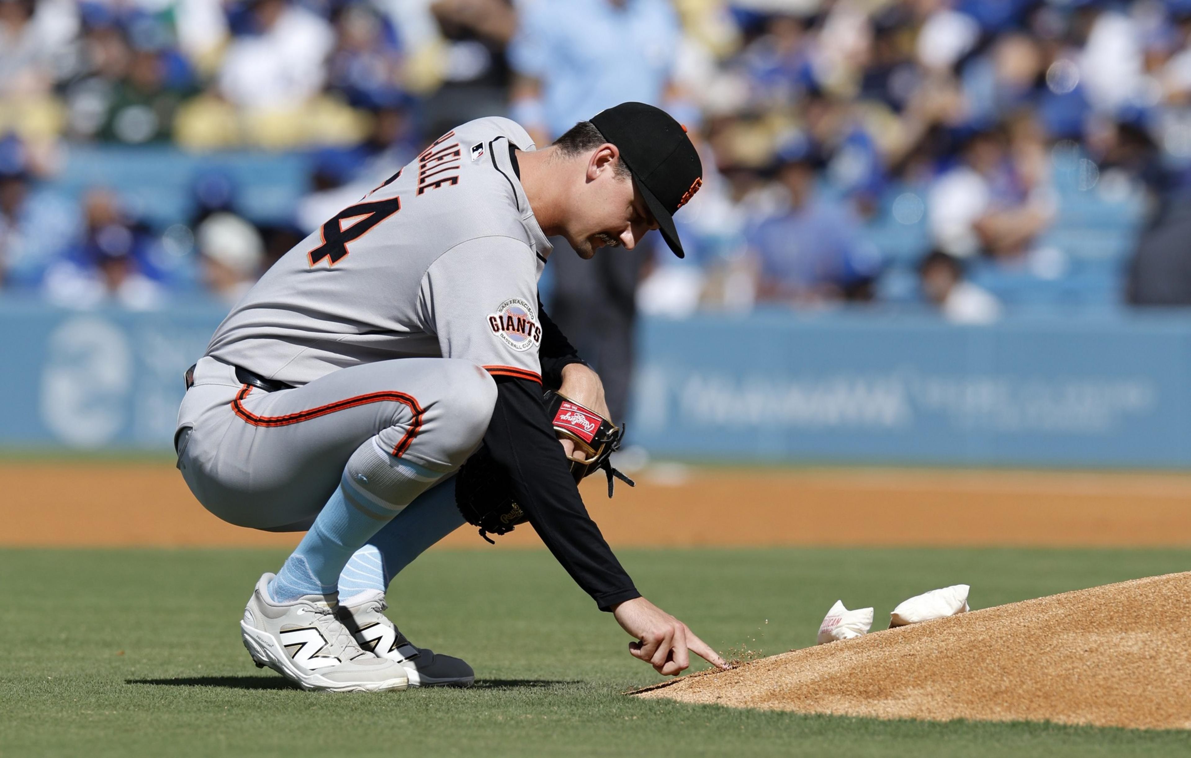 A baseball player in a gray uniform crouches near the pitcher's mound, touching the ground. He wears a black cap, and the crowd is blurred in the background.