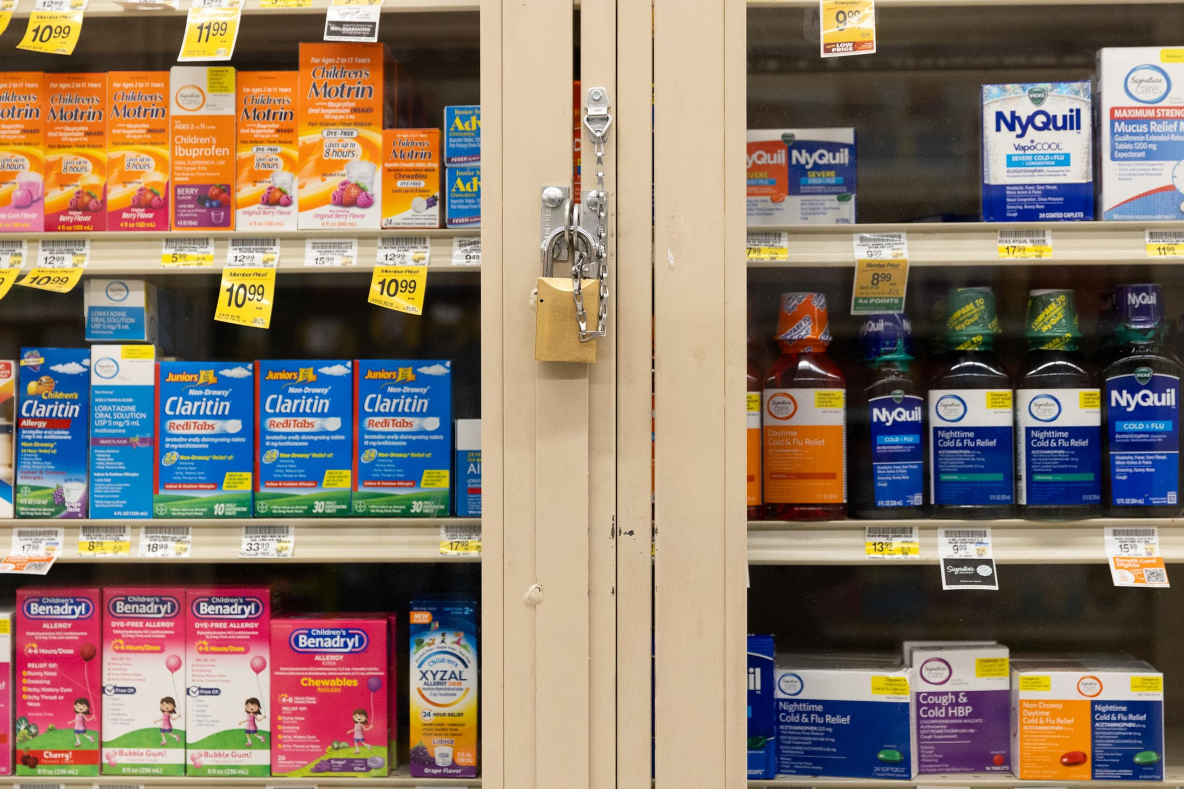 The image shows a store shelf with various cold and allergy medications like Children's Motrin, Claritin, and NyQuil, secured in a glass cabinet with a padlock.